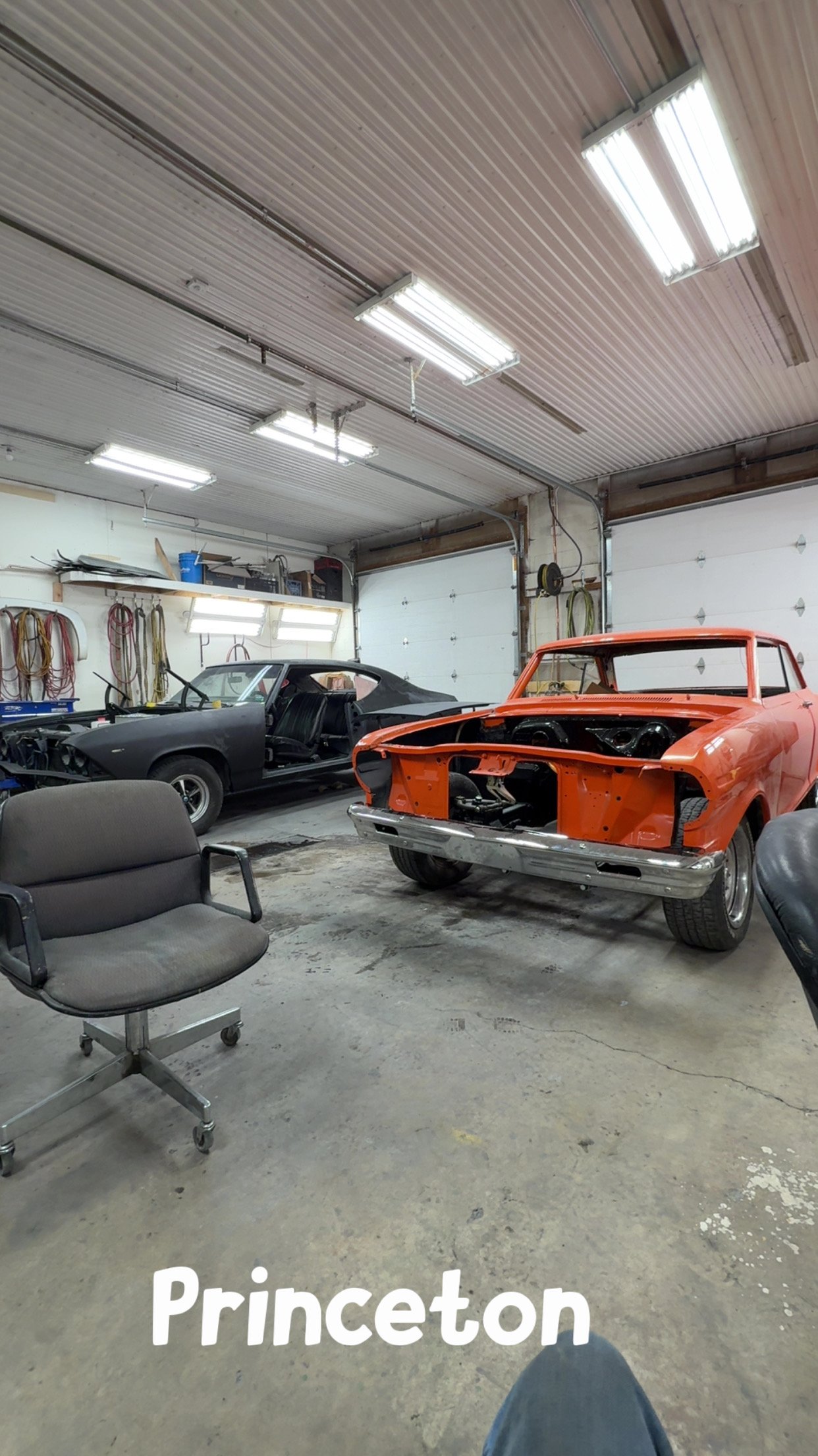 A workshop with two cars under restoration, one red and one black, and various tools hanging on the wall. The word 'Princeton' is visible at the bottom of the image.
