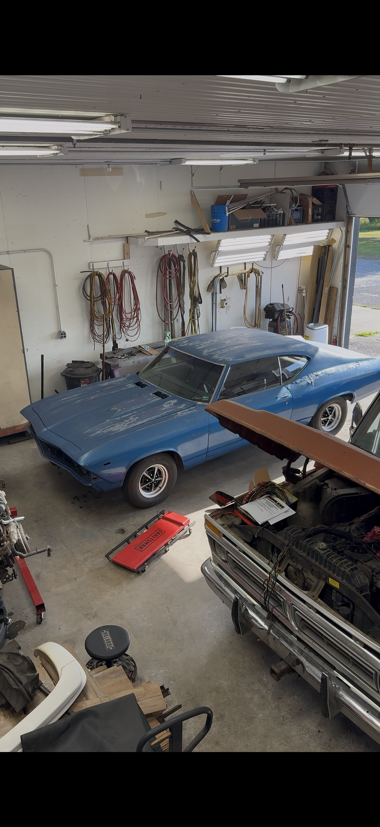 A blue vintage car parked inside a cluttered garage with various tools, wires, and equipment hanging on the walls and stored on shelves.