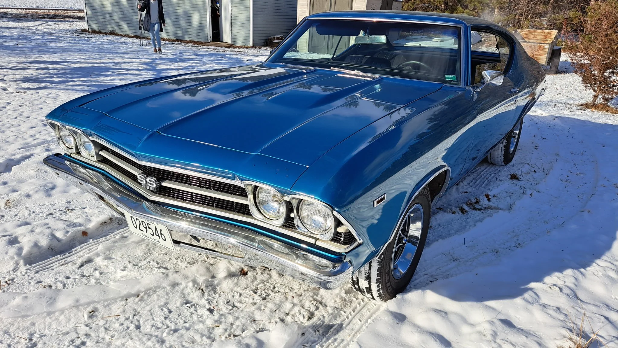 A vintage blue Chevrolet SS car parked outside on snow-covered ground near a building with a person in the background.