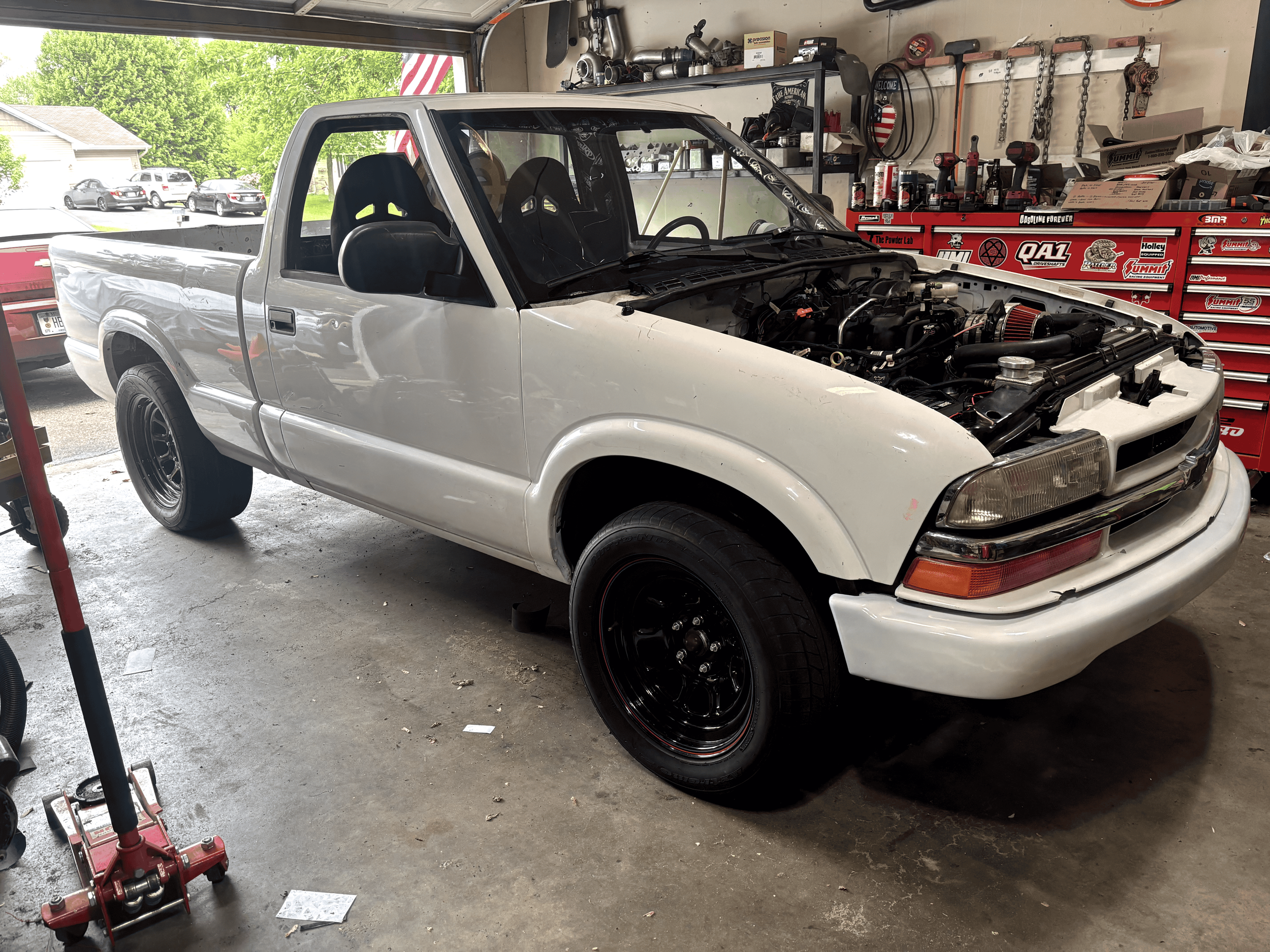 A white pickup truck is being worked on inside a garage. The truck has an open hood with visible engine parts. The garage has a red tool chest, shelves with various tools and car parts, and an American flag hanging on the wall. Outside the garage, parked cars and green trees are visible.
