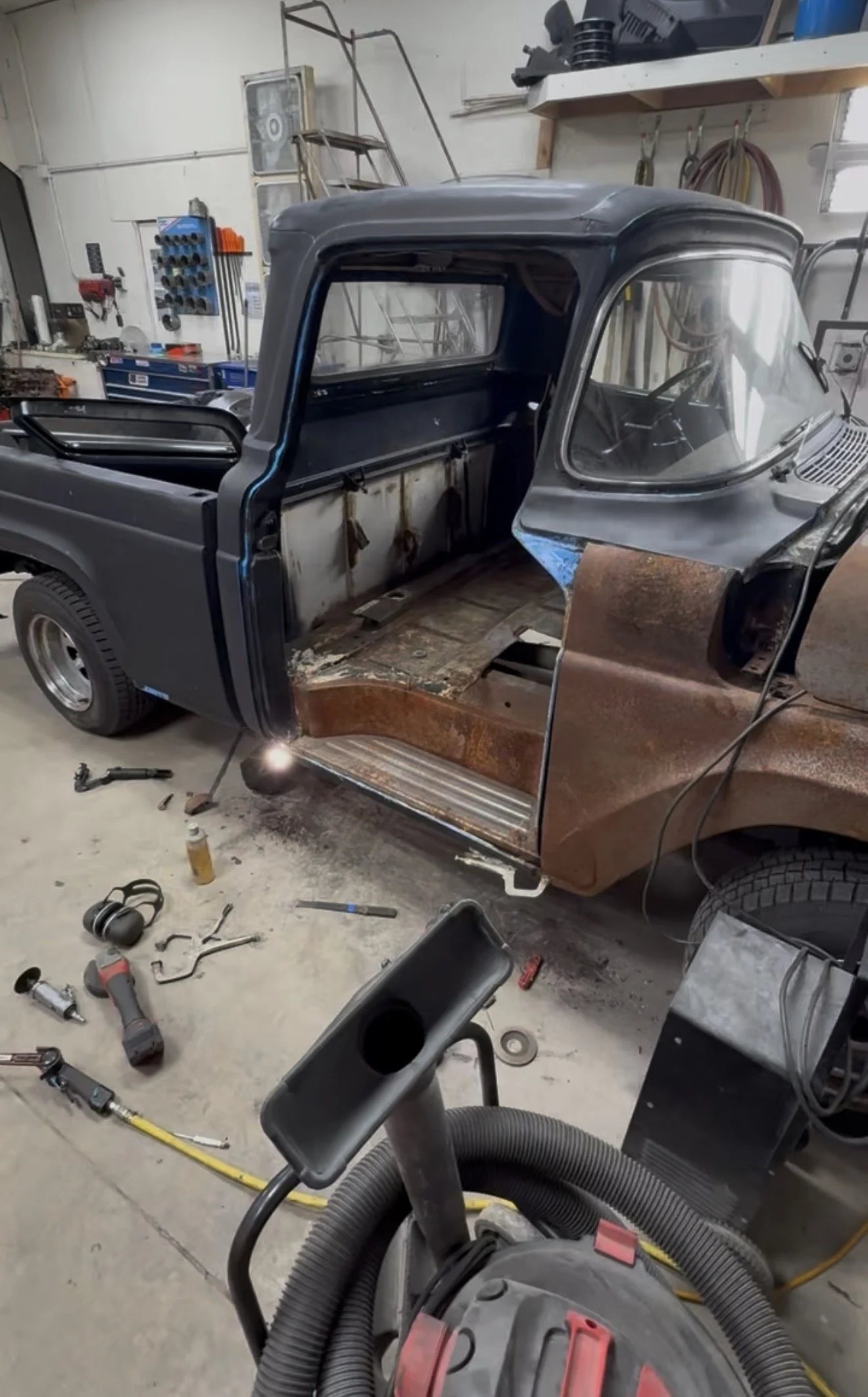 Disassembled vintage vehicle truck cab in a workshop with tools, equipment, and rusted metal parts scattered around.