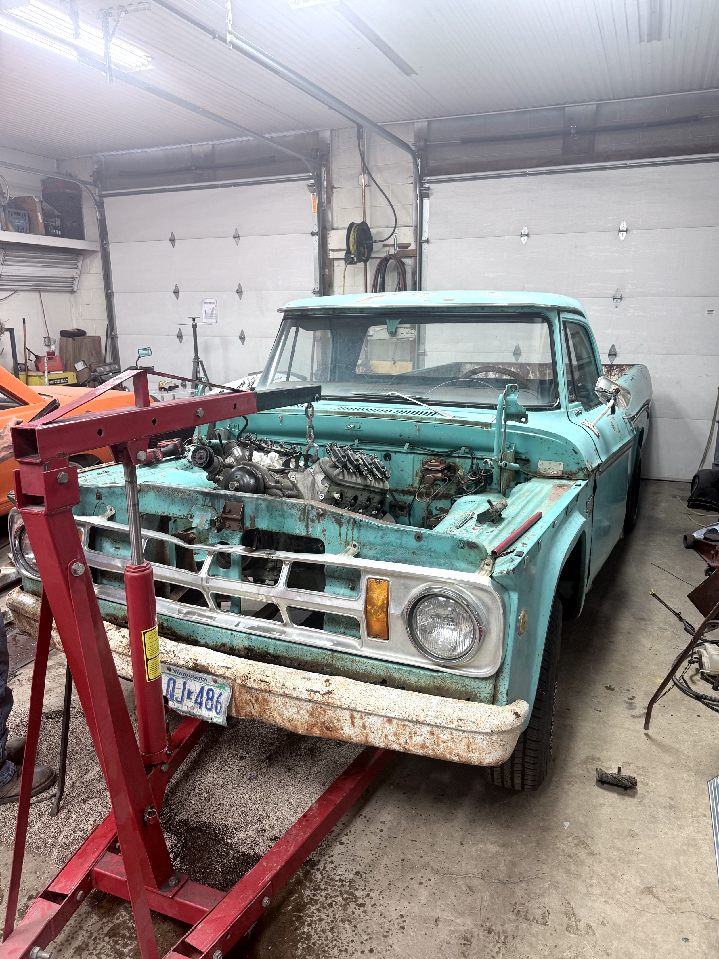 An old, rusted light blue pickup truck inside a garage, with a red engine hoist in front of it. The truck appears to be under repair or restoration, with its hood removed. Garage walls have various tools and equipment hanging, and the floor is concrete with some oil stains.