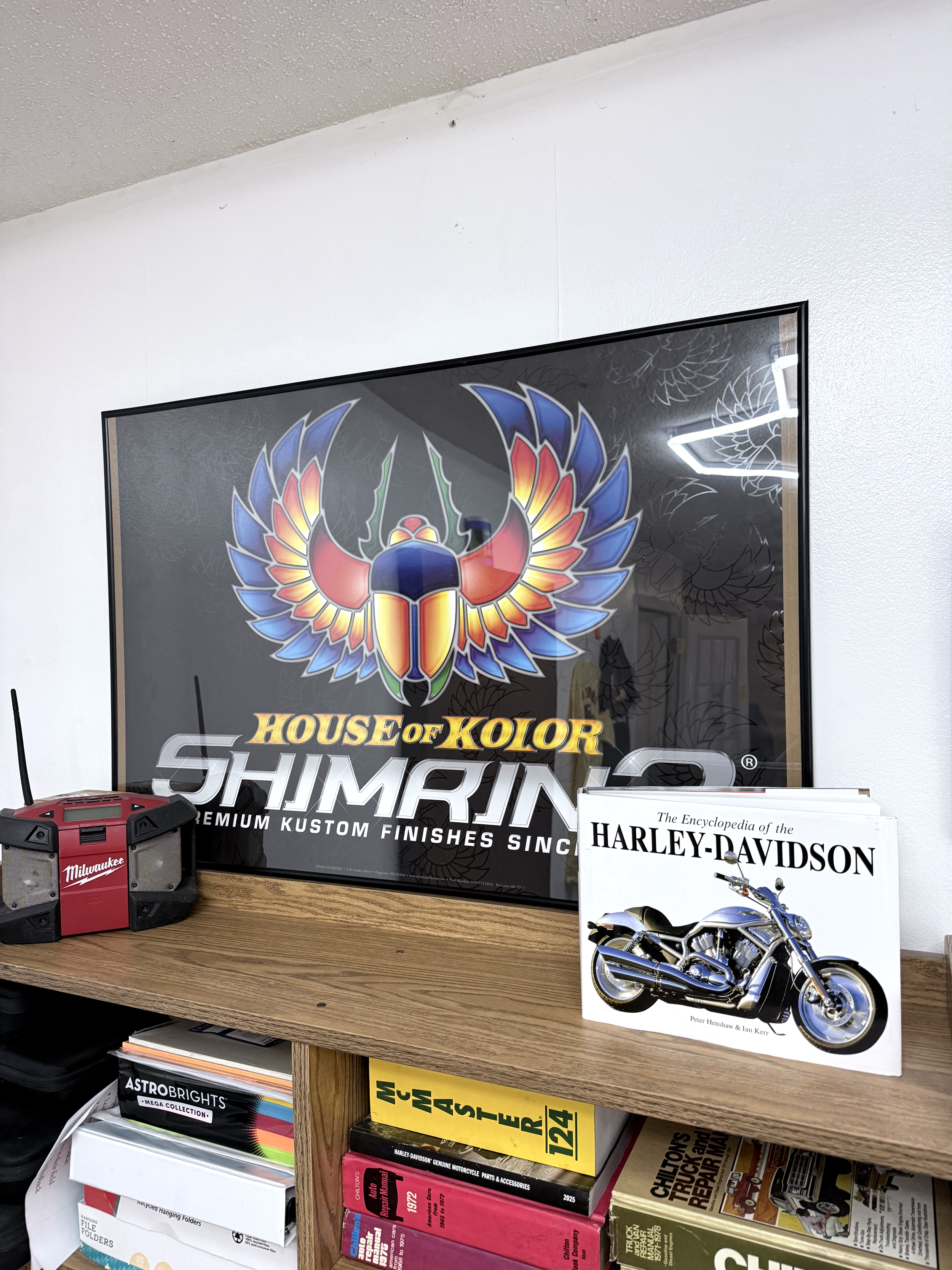 A wooden table with a Milwaukee portable speaker, a Harley-Davidson book titled "The Encyclopedia of the Harley-Davidson," a motorcycle model, and a bookshelf with various books including "Astro Brights" and "Trucks & Truck Repair."