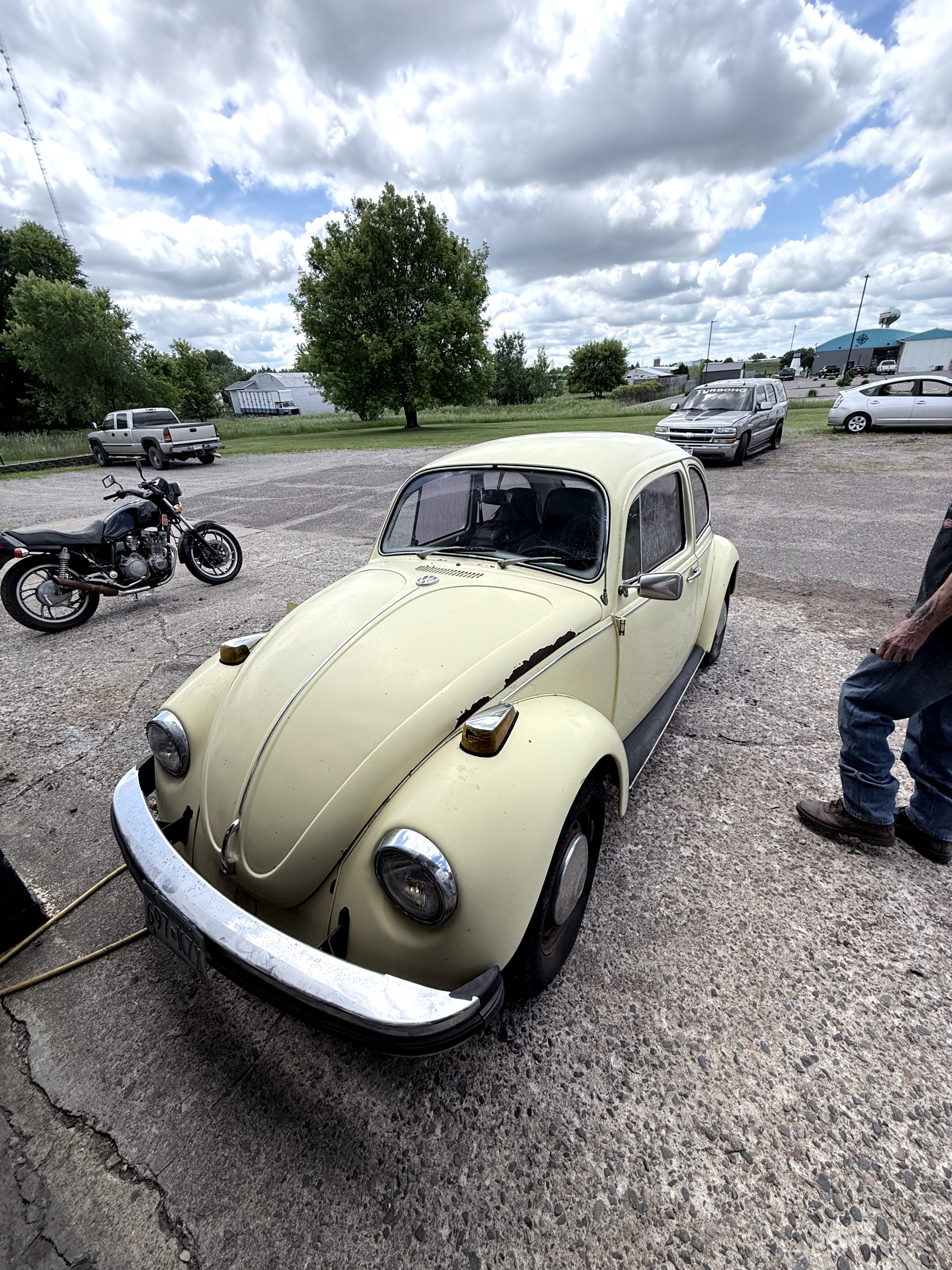 A pale yellow, vintage Citroën 2CV car parked on a gravel surface in a lot with other vehicles, including a motorcycle, pickup truck, and modern cars, under a cloudy sky with trees and buildings in the background.