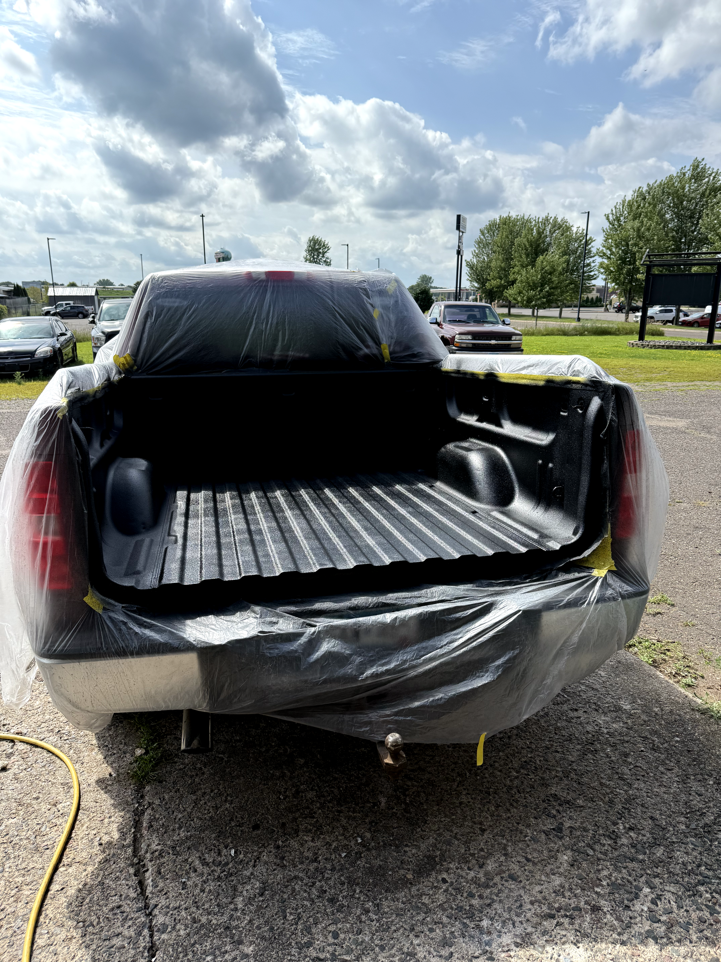 The image shows the back of a pickup truck that has been covered with plastic and painter's tape, likely in preparation for painting or repair work. The bed of the truck is empty, and the truck is parked outdoors on a paved surface with a grassy area and several other parked cars in the background, under a partly cloudy sky.