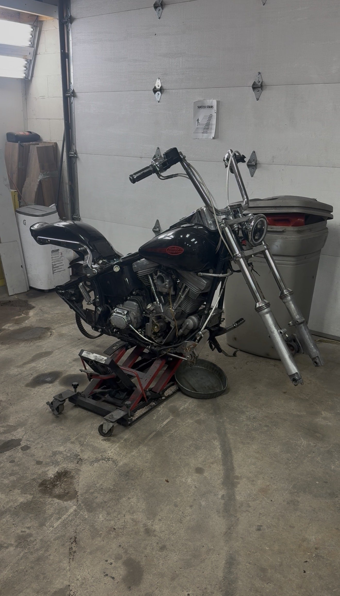 A motorcycle being repaired in a garage, with some parts removed and placed on a roller stand. The garage has a concrete floor, a closed garage door, some storage items, and a garbage can.