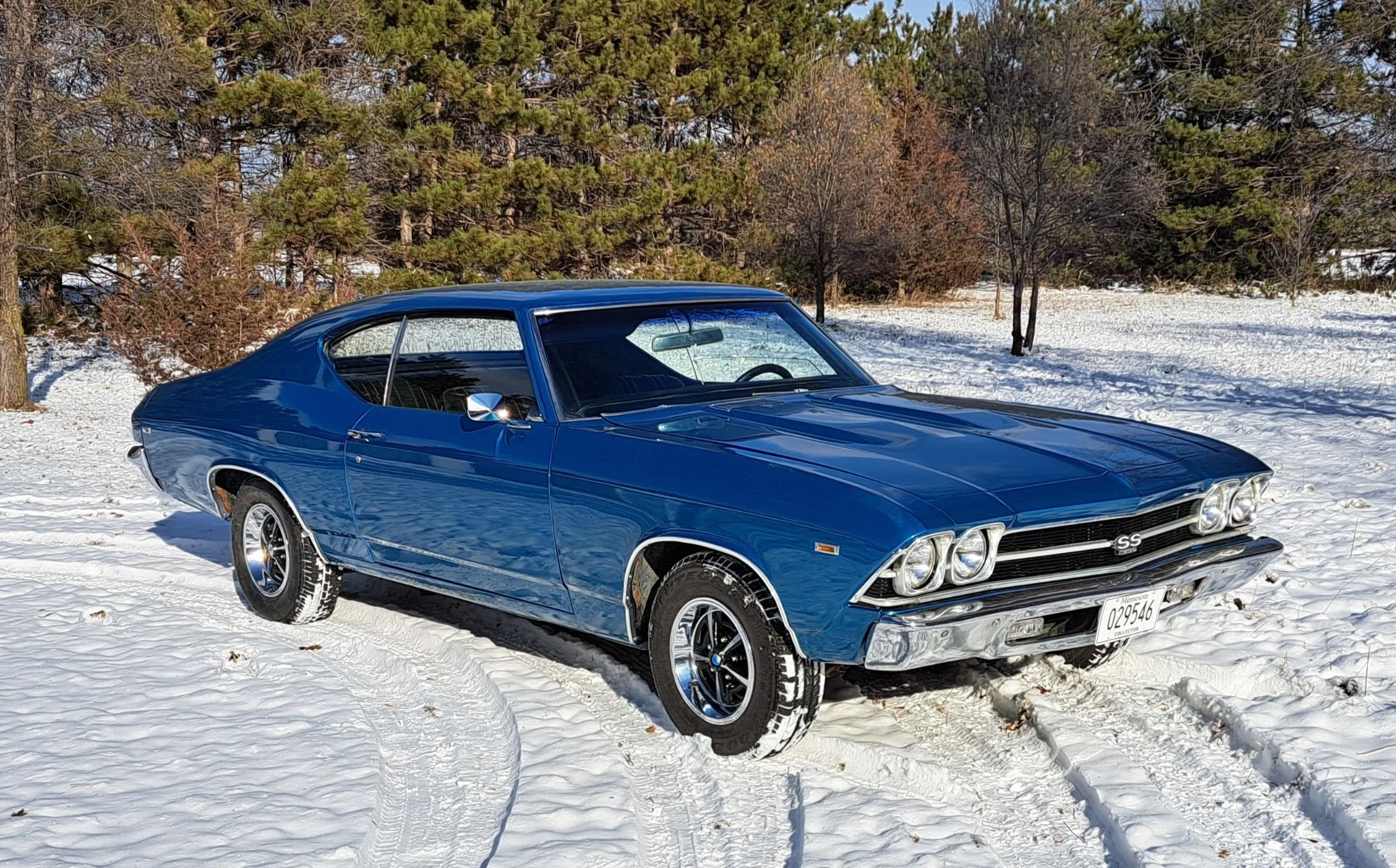 Blue vintage Chevrolet Chevelle SS parked on snowy ground with pine trees and leafless trees in the background.