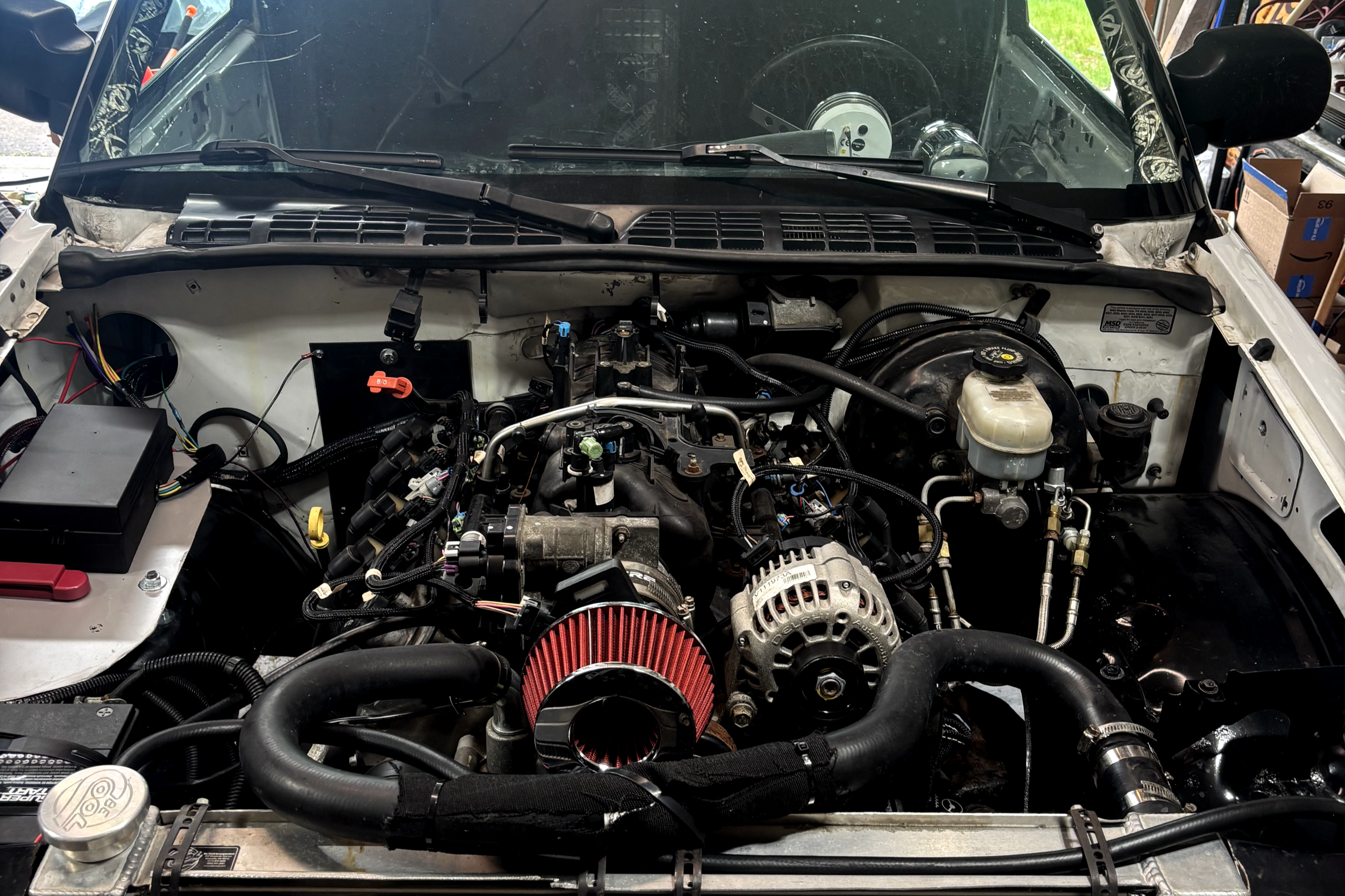 View of a car engine with a red air filter, various hoses, wires, and mechanical components visible inside the engine bay.