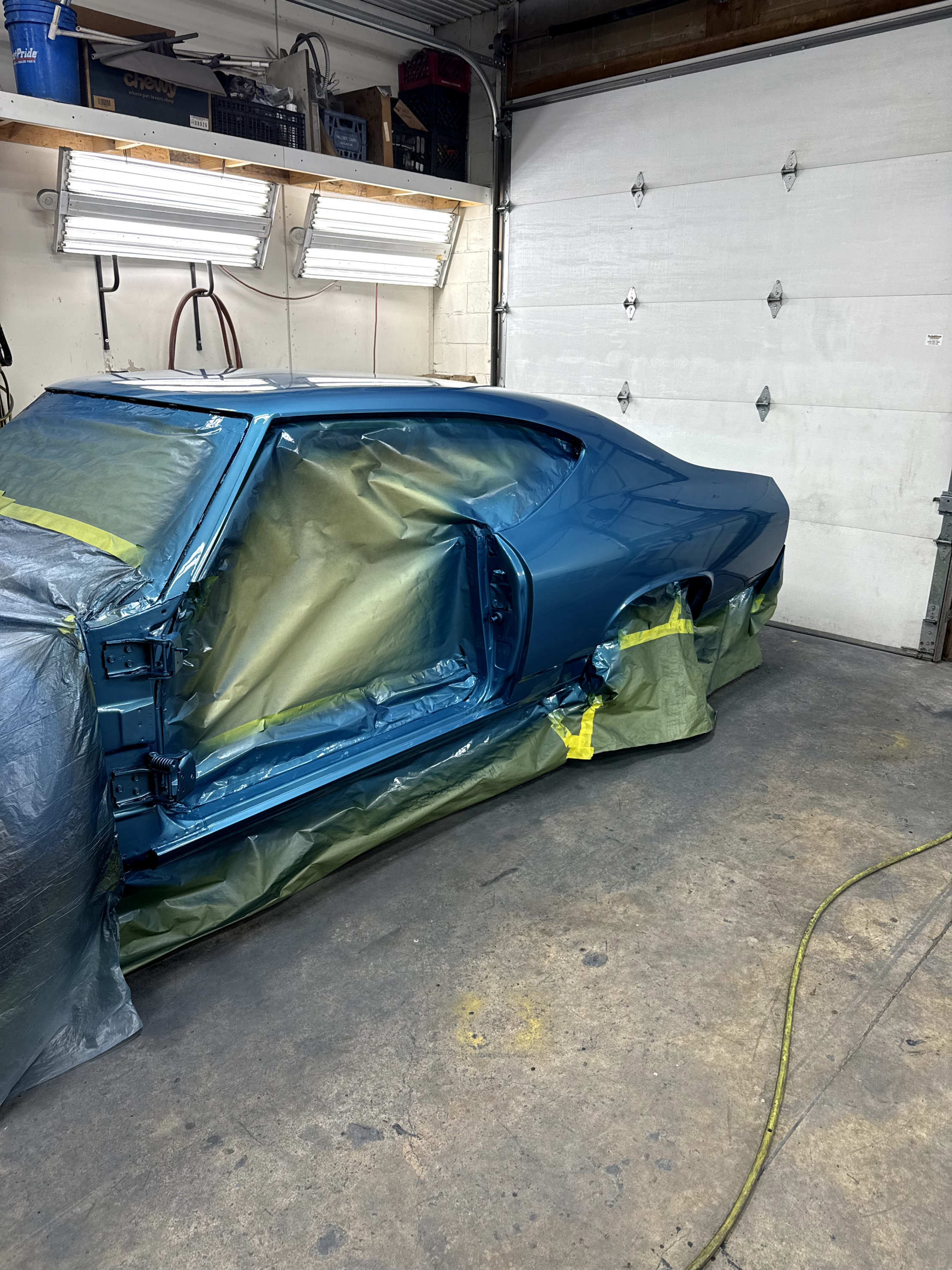 A car with a blue paint job is in a garage, covered partially with masking paper and taped, likely for painting or repair. The garage has a concrete floor, a large white door, and fluorescent lighting.