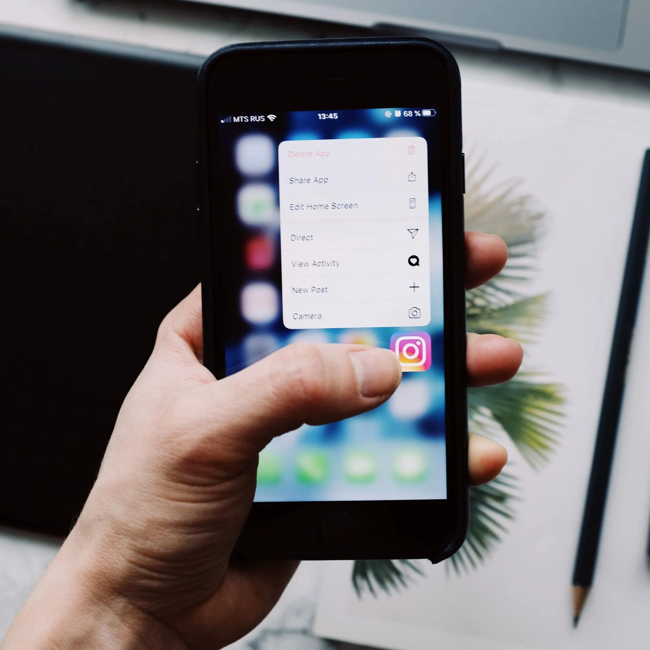 Close-up of a smartphone screen displaying options like Delete App, Share App, Edit Home Screen, Direct, View Activity, New Post, and Camera. The phone is held by a person's hand, with a blurry background including a plant and a gray surface.