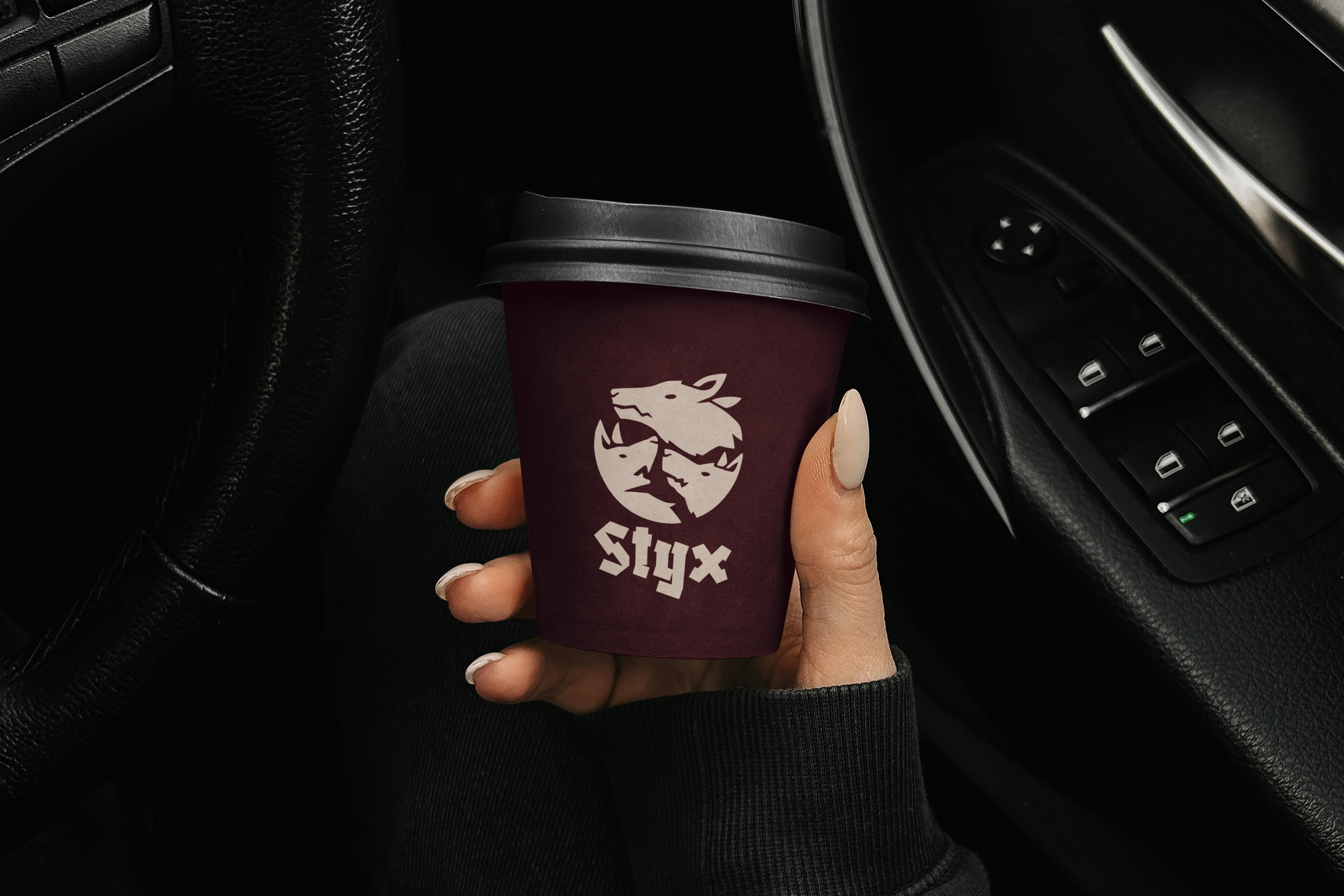 Person holding a maroon paper coffee cup with a lion logo and the words 'FELIX STYX' inside a car, near the steering wheel and dashboard.