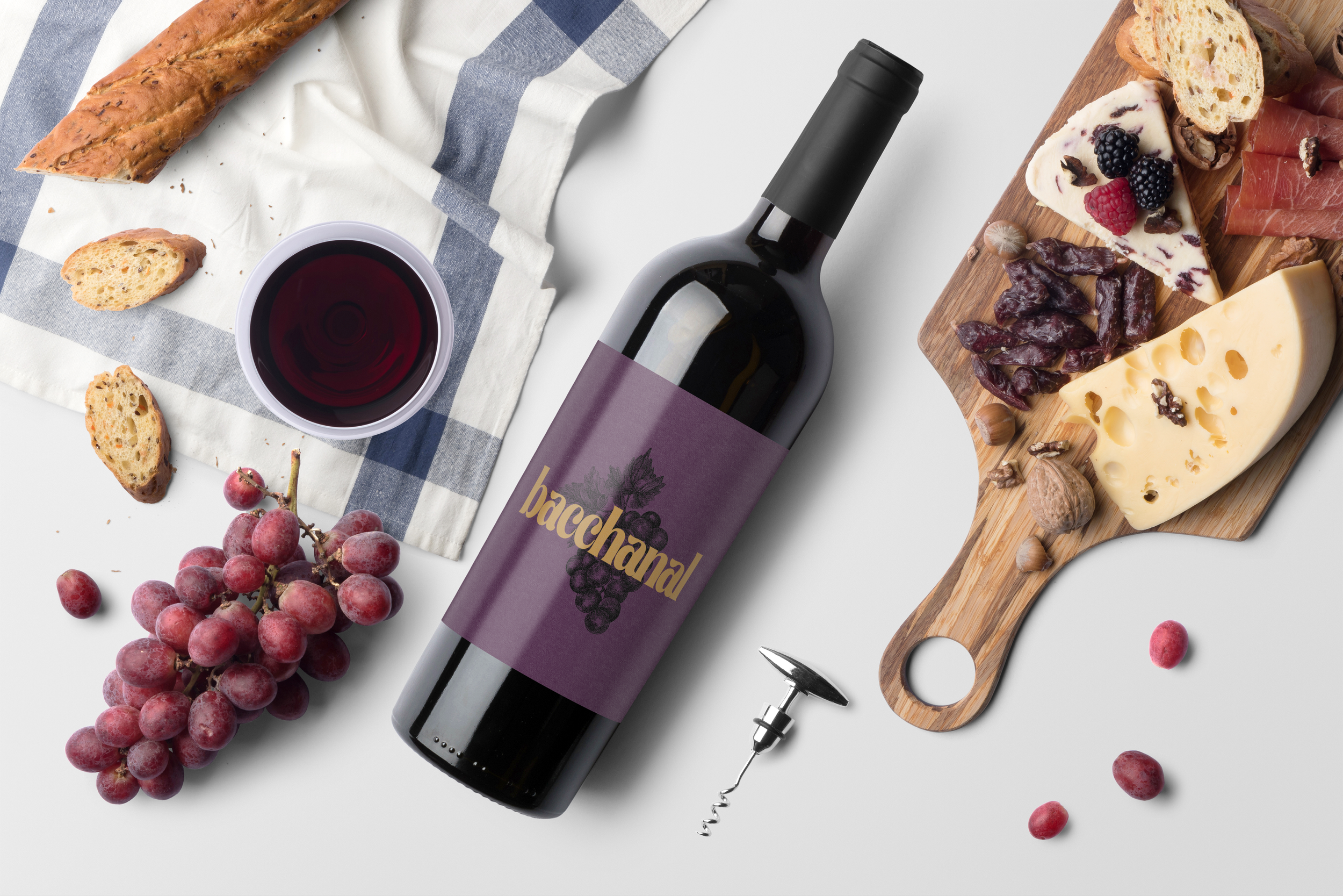 A bottle of red wine, a bunch of red grapes, a cheese board with various cheeses, slices of bread, and charcuterie. A glass of red wine, a corkscrew, and a white and blue checkered cloth are also present on a white surface.