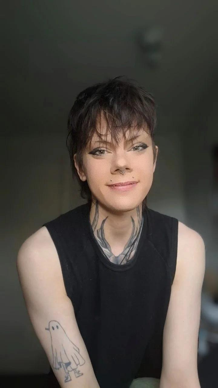 A young woman with short, dark hair, wearing a black sleeveless top, has tattoos visible on her neck and arm, and is smiling at the camera.