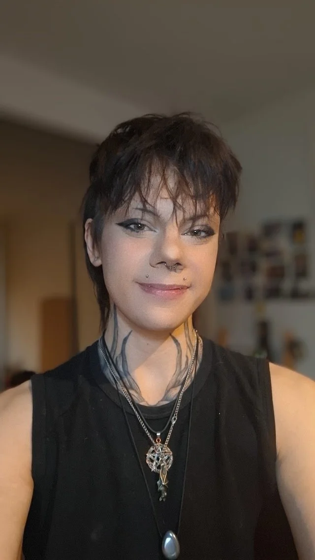 A person with short, dark, tousled hair smiling at the camera, wearing black eyeliner, multiple necklaces including a large pendant, a septum piercing, and tattoos on their neck and chest, in a room with blurred background.