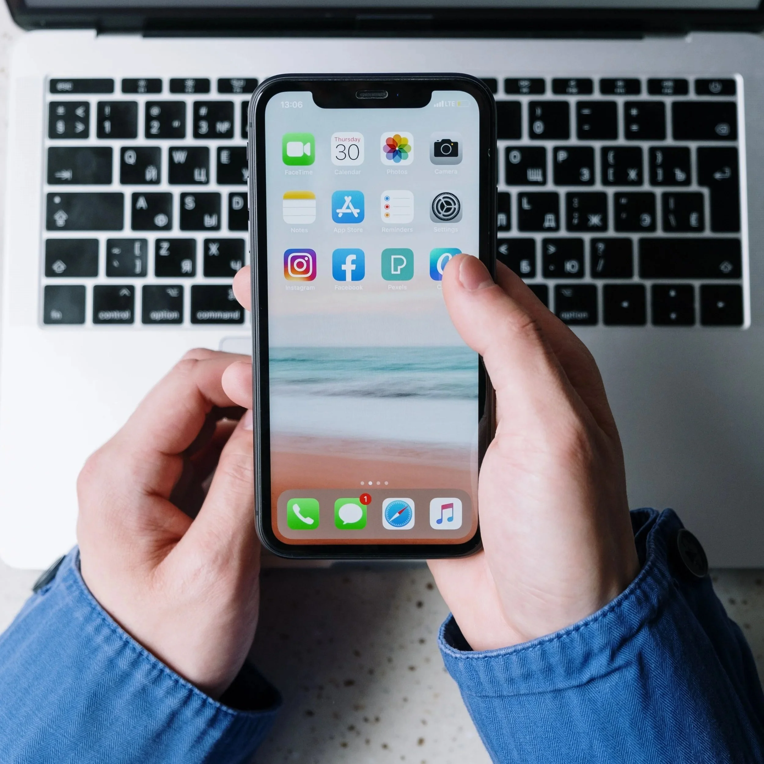Person holding a smartphone over a laptop keyboard, showing app icons on the phone screen.