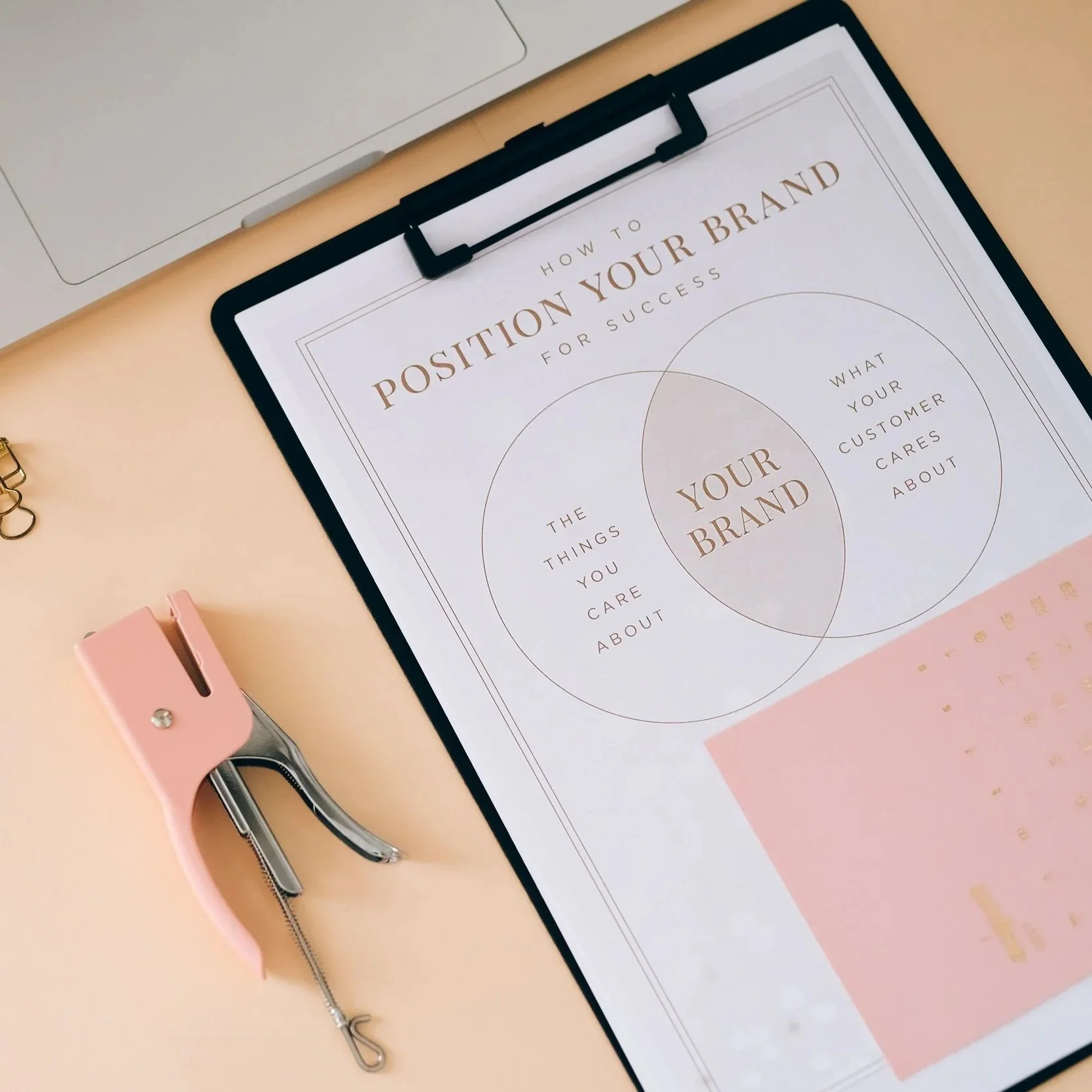 A flat lay photo of a clipboard with a printed document titled "How to Position Your Brand for Success" and a Venn diagram, a pink paper clip, a pink binder clip, and a silver stapler on a pink surface.