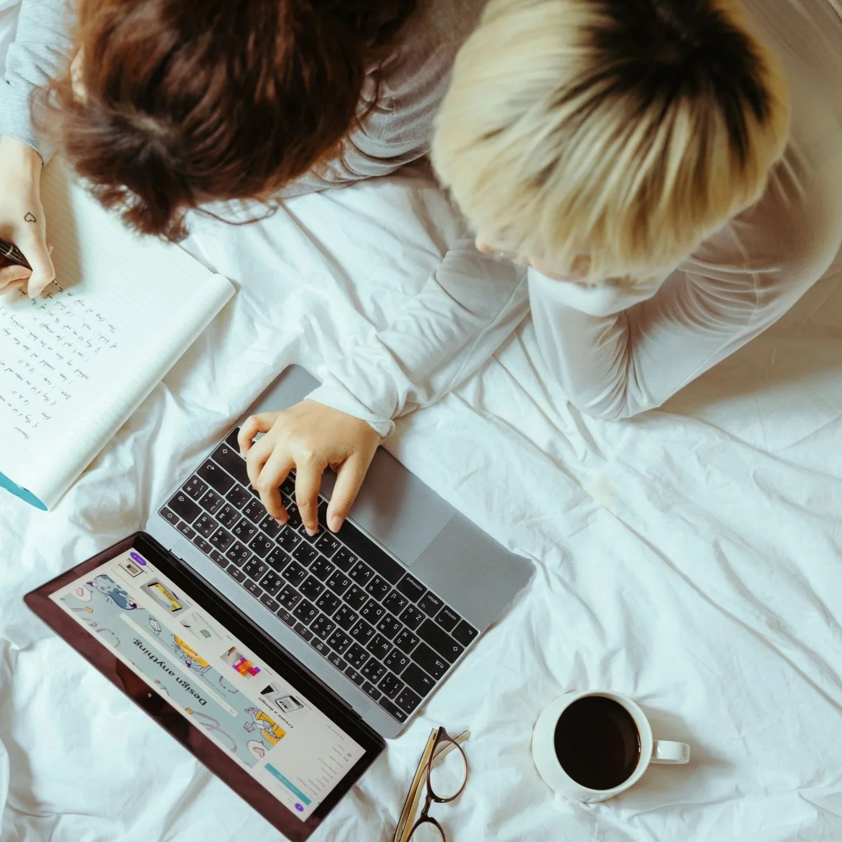Overhead view of a person with blonde hair working on a laptop, with a phone displaying a website, a cup of coffee, a pair of glasses, a notebook, and a pen on a white bed.