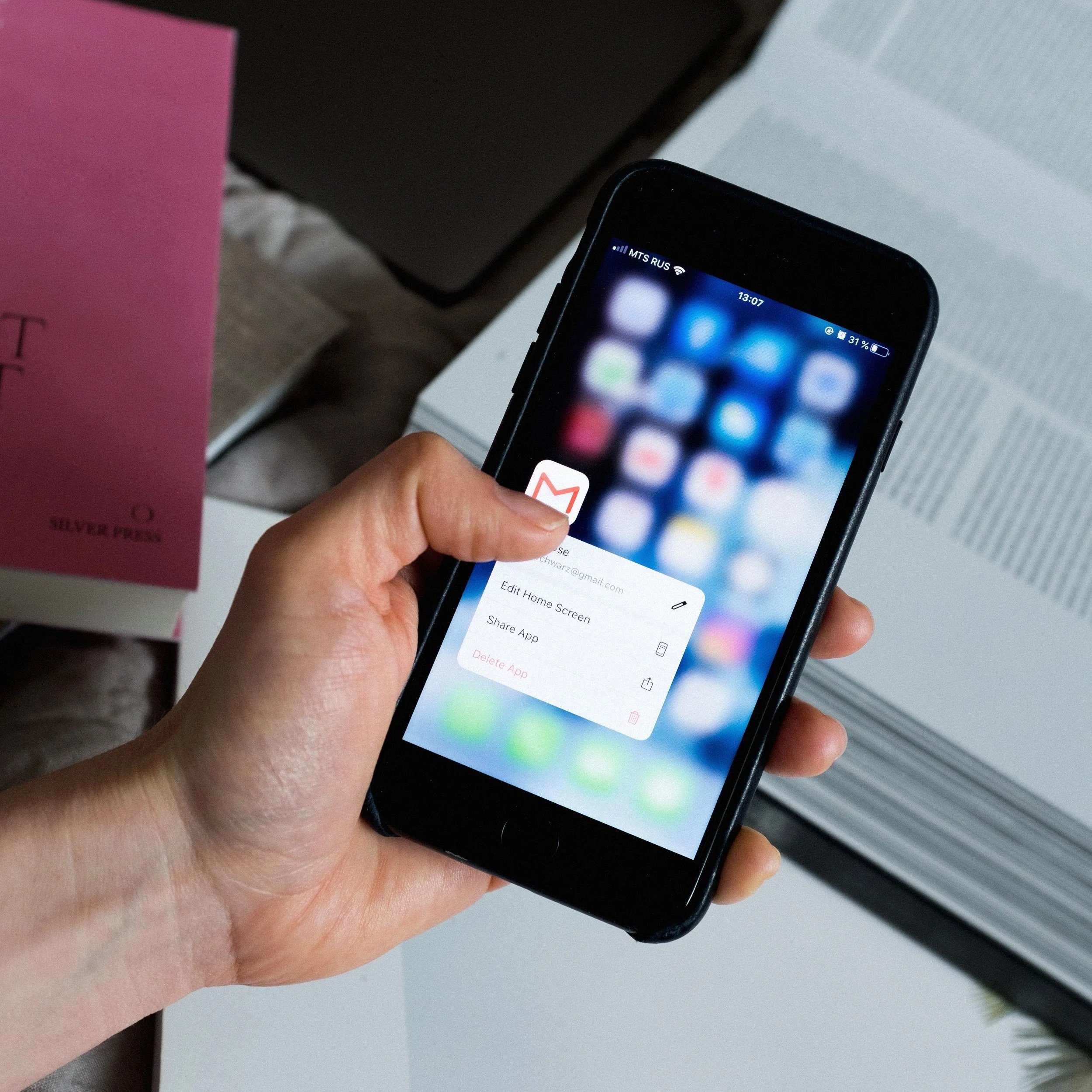 Person holding a smartphone showing app menu, surrounded by books and a pink box.