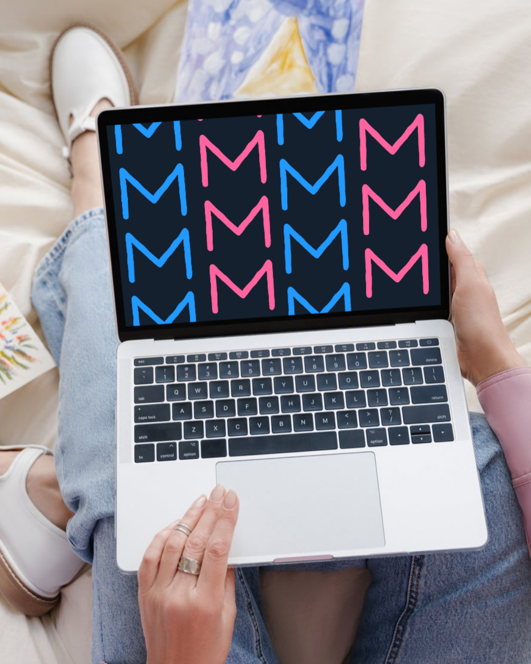 A person sitting on a beige bed holding a laptop with a colorful pattern of pink and blue 'M' shapes on the screen.