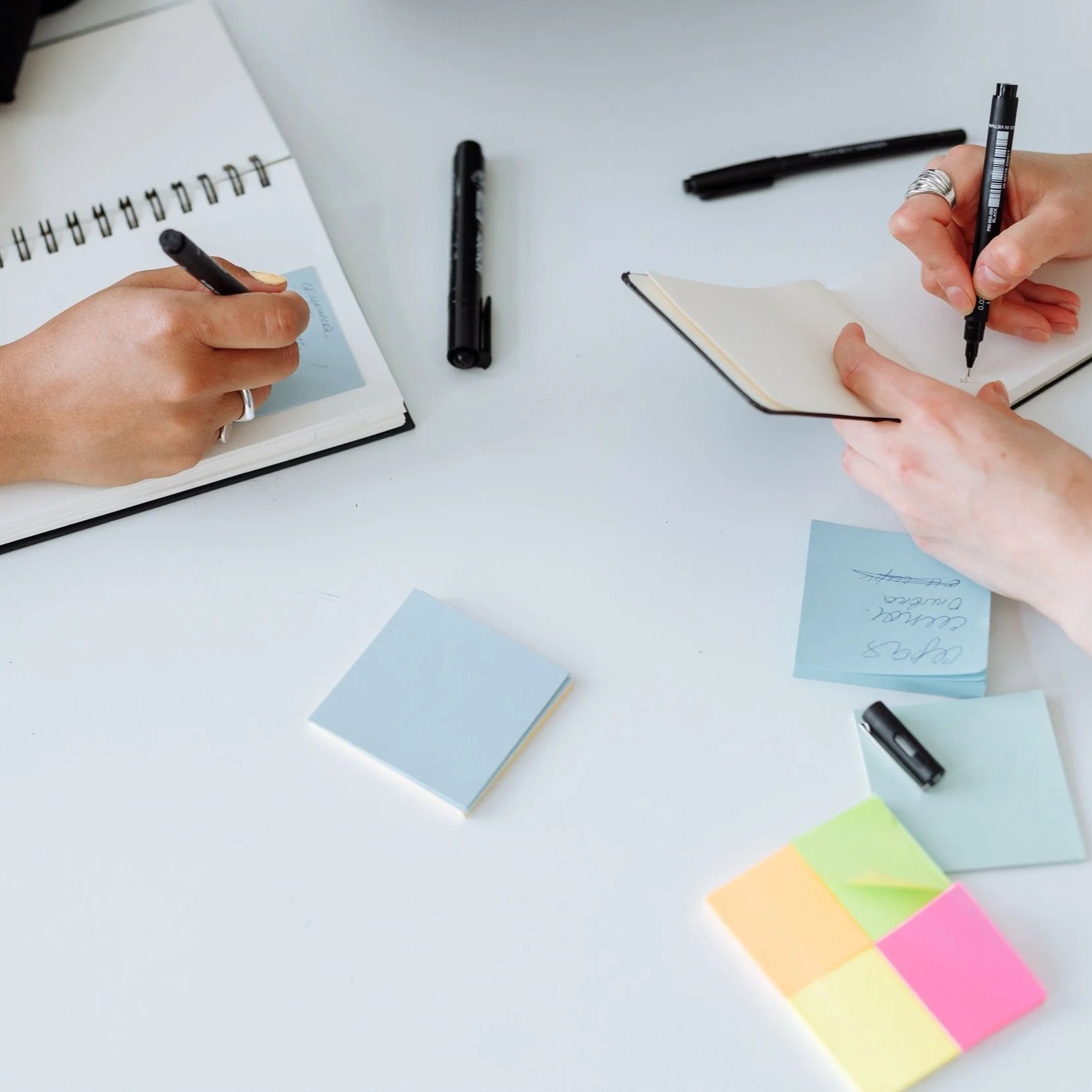 Hands writing in notebooks and note papers on a white desk, surrounded by markers and colorful sticky notes.