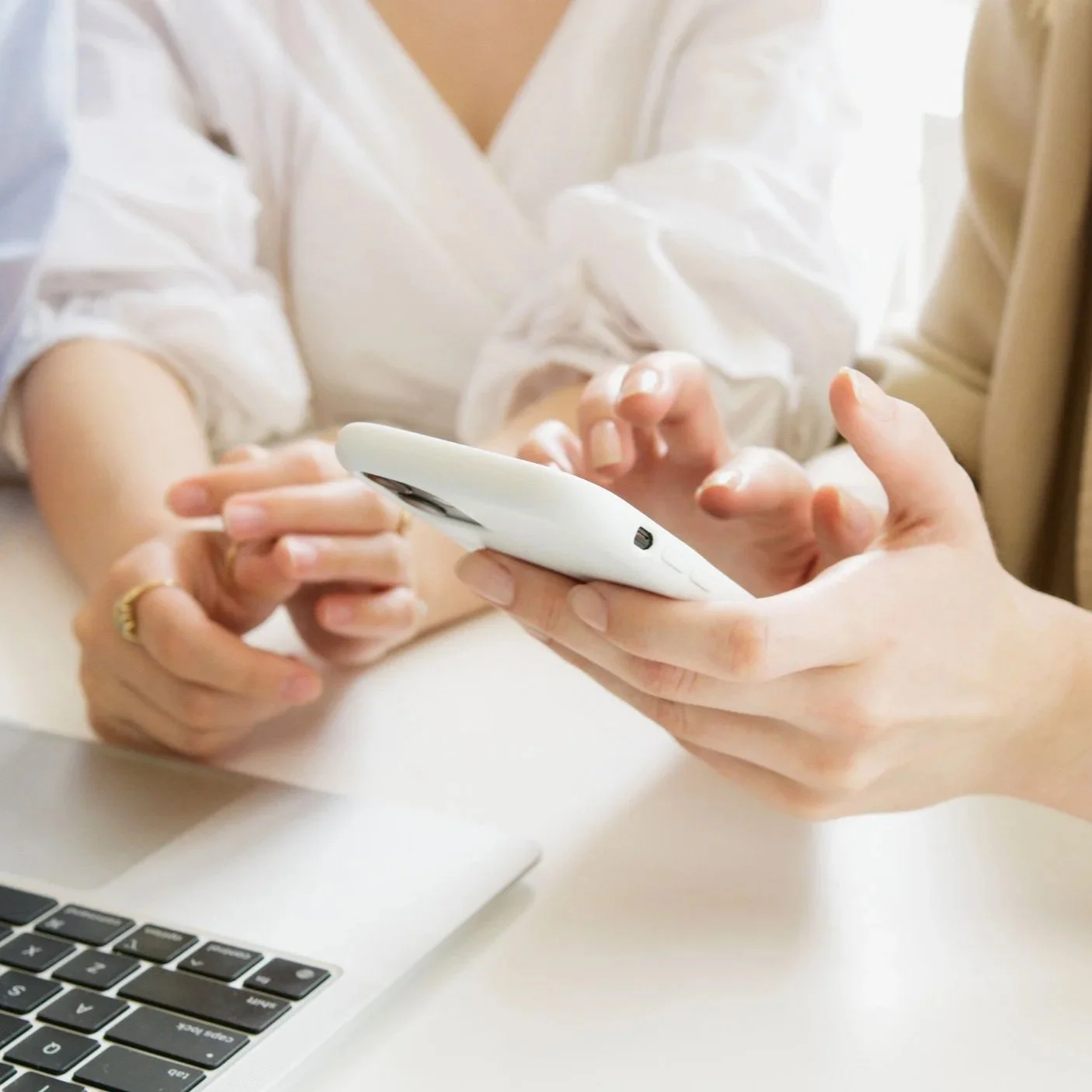 Two people using a white smartphone collaboratively, with a laptop nearby on a white table.