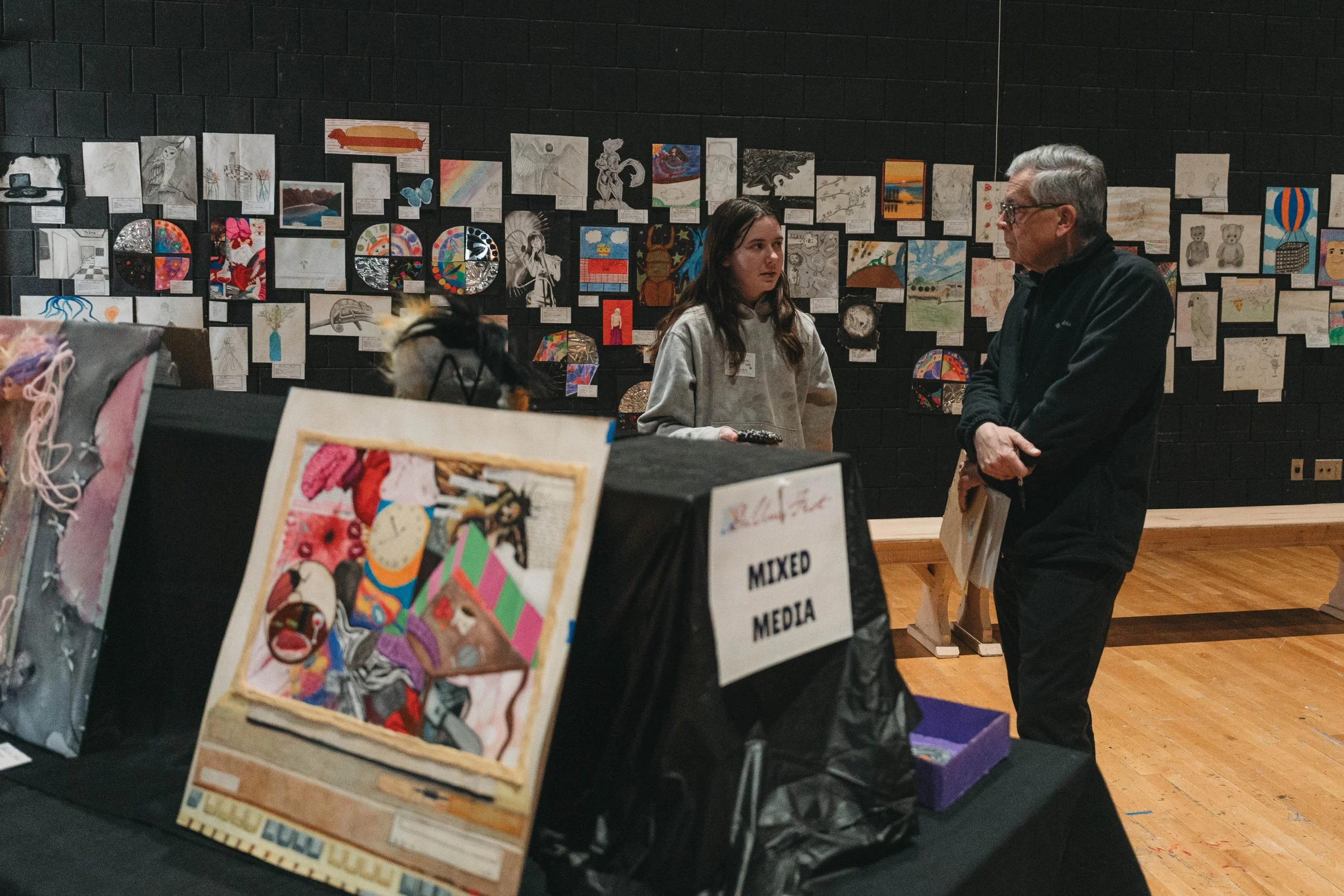View of an art exhibition with artworks on a black wall, two people in conversation, and a table displaying various art pieces.