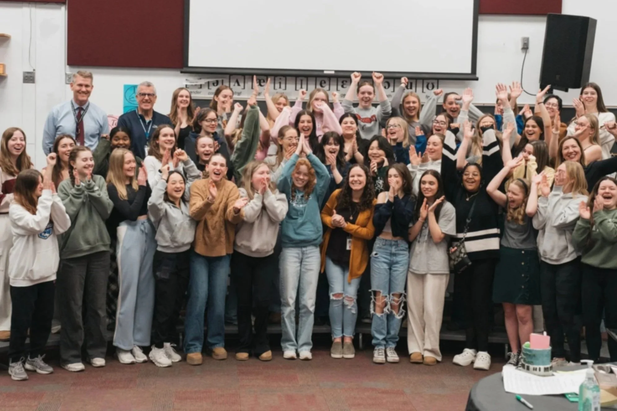 Group of smiling students and teachers celebrating together in a classroom with a whiteboard and projection screen behind them.
