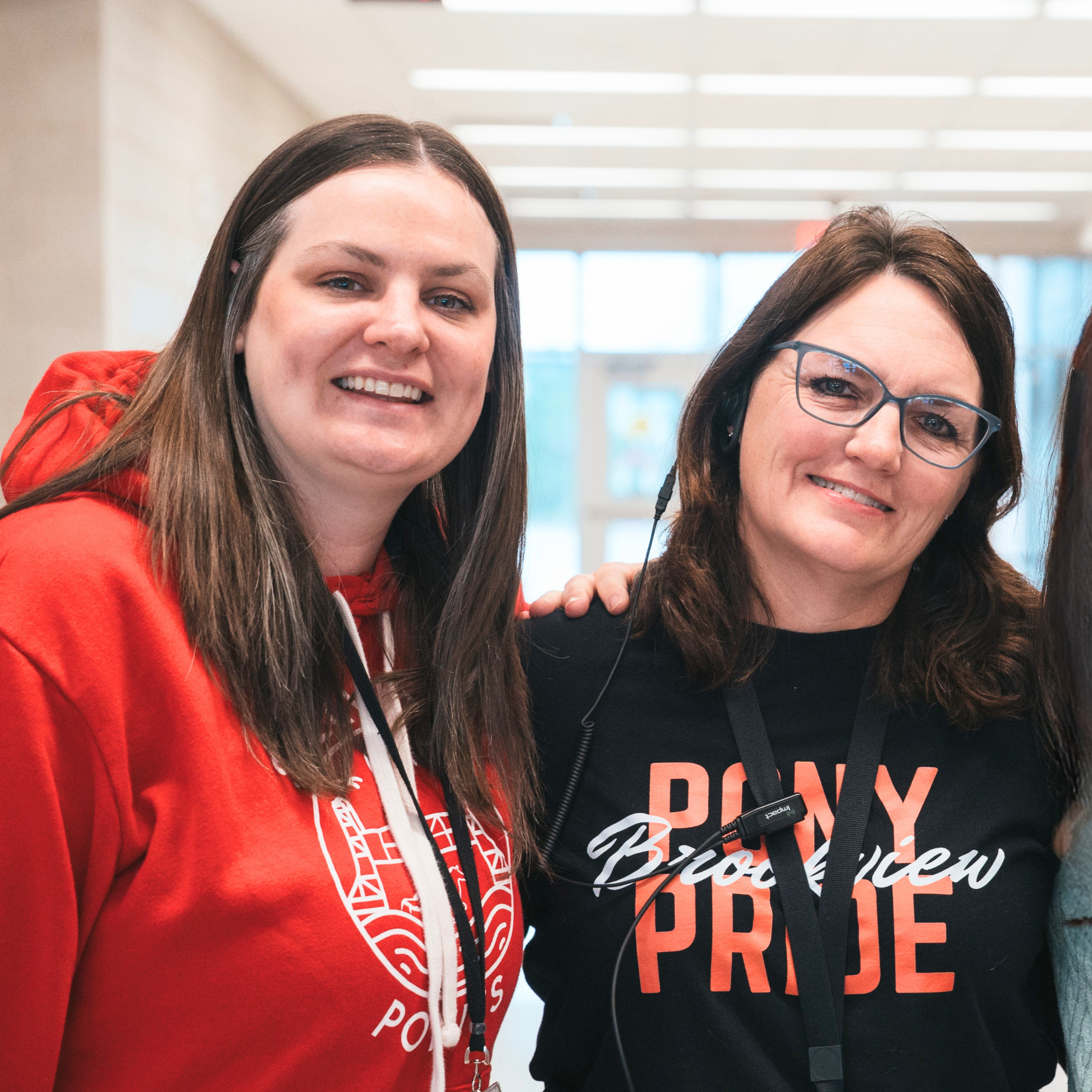 Close-up of two women smiling, one wearing a red hoodie with a white design and the other wearing glasses and a black shirt with orange text, indoors with bright lighting.