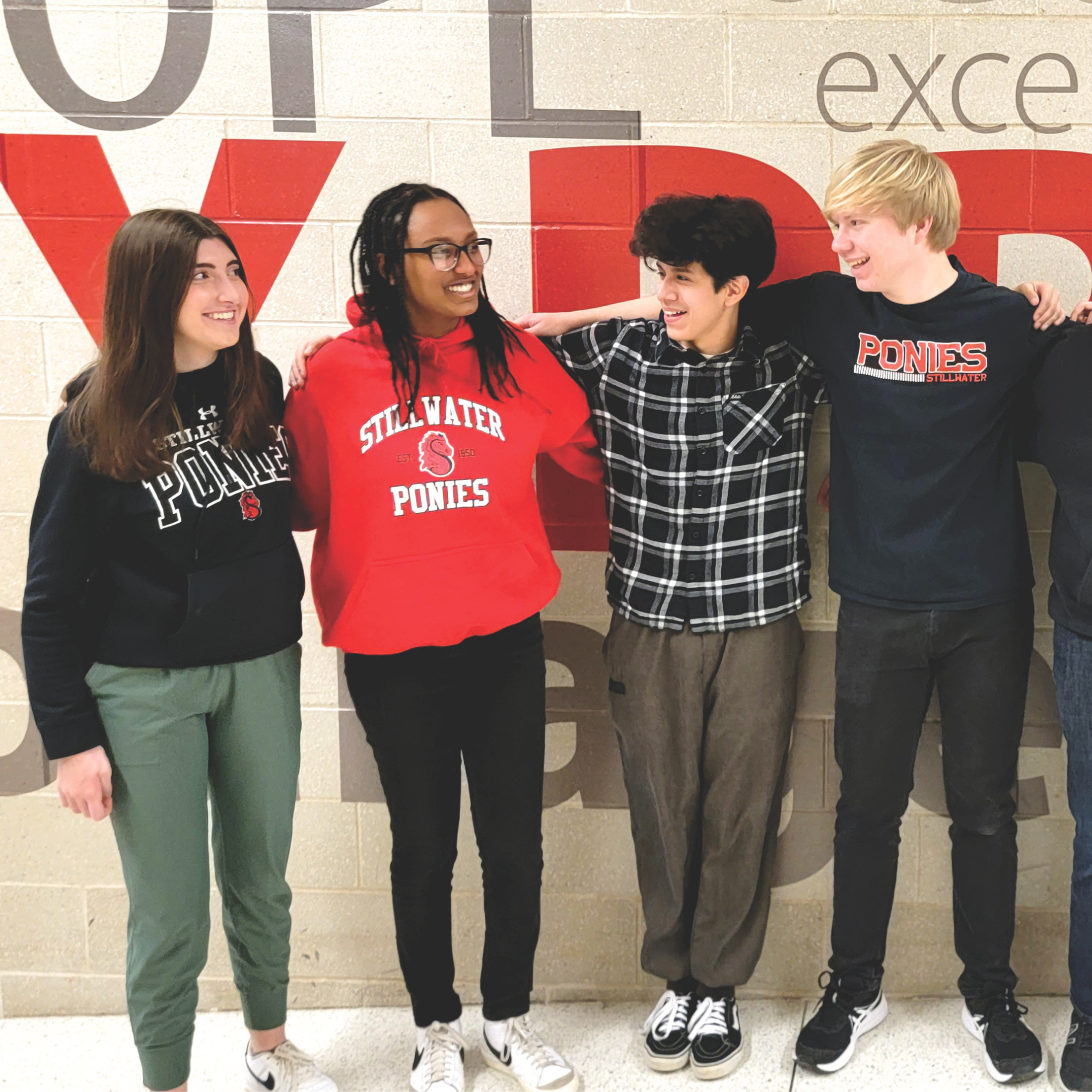 Four teenagers, two girls and two boys, standing side by side against a beige tiled wall with red and gray text, smiling and having a conversation, wearing casual clothing with sports team logos.