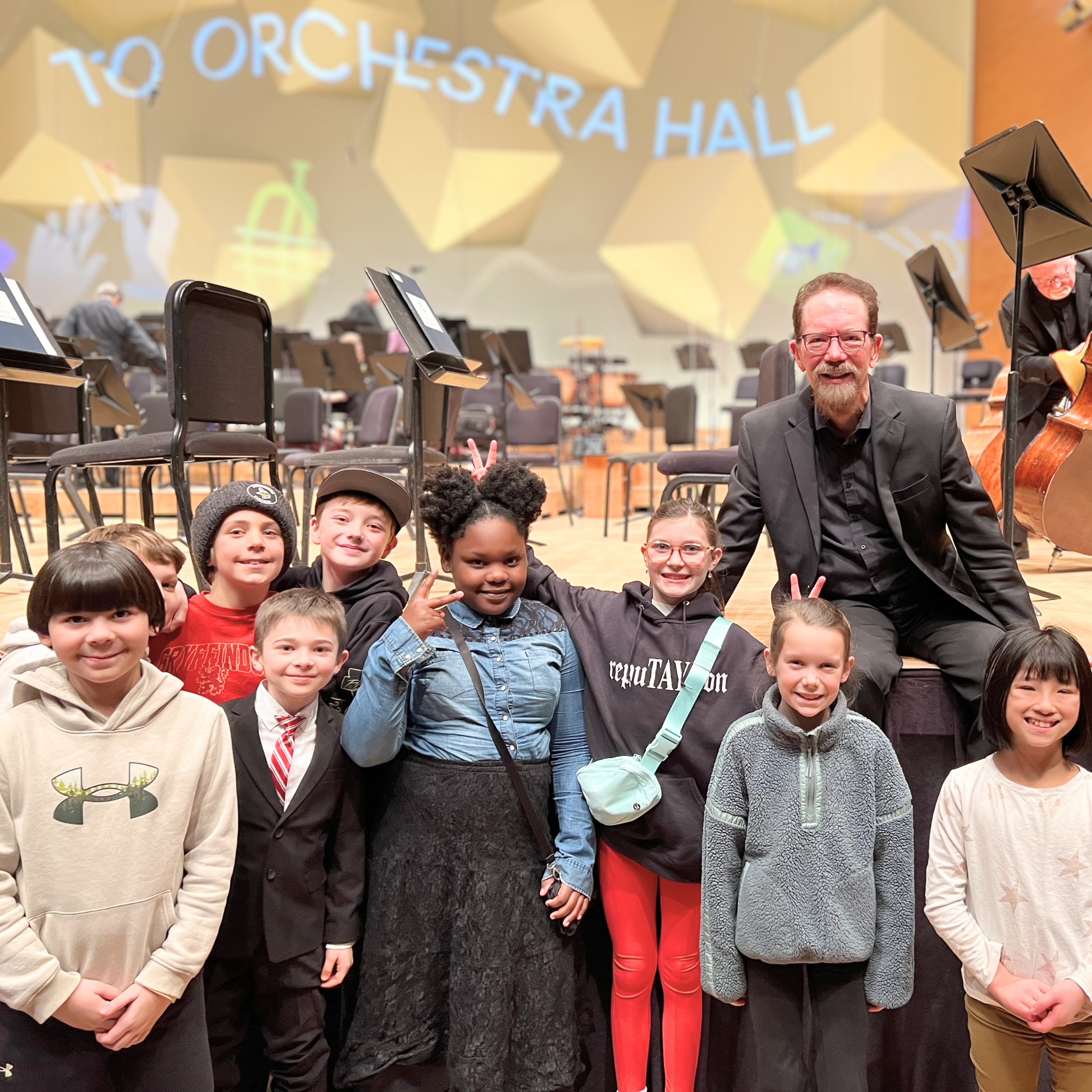 Group of children and a man with a cello posing in an auditorium with a stage, chairs, music stands, and a large screen in the background displaying the words 'TO ORCHESTRA HALL'.