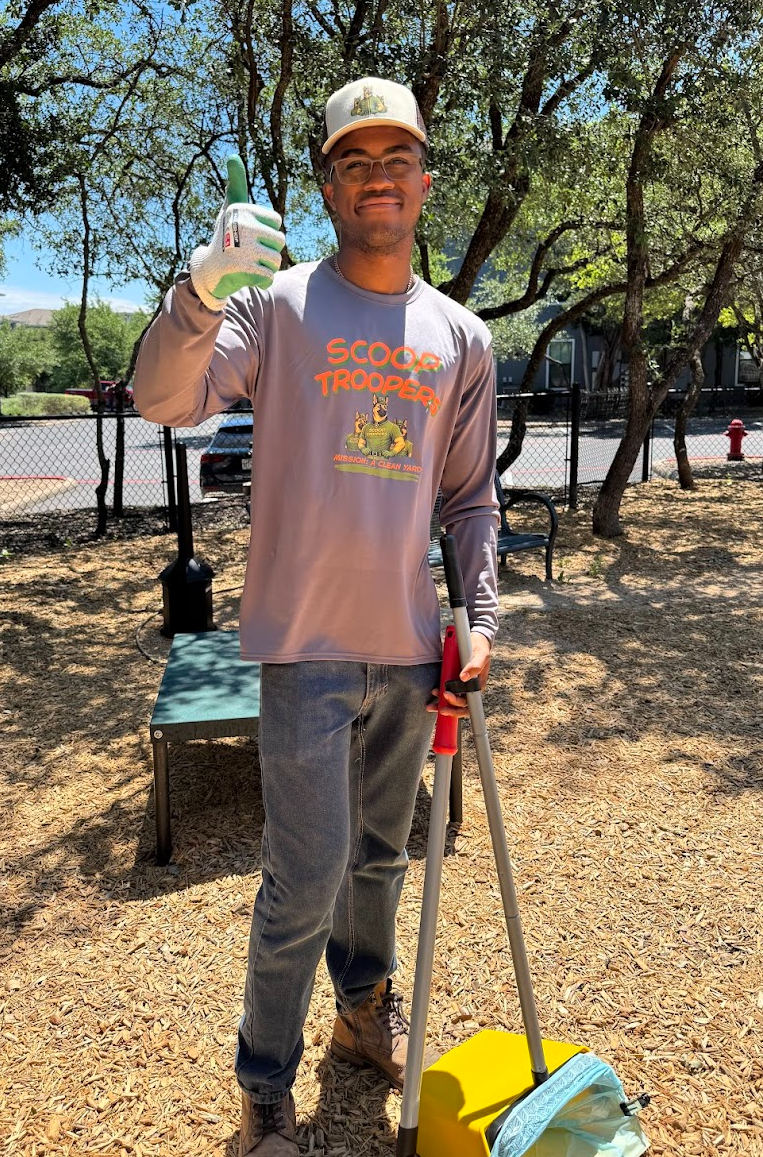 A young man wearing glasses, a baseball cap, and a long-sleeve shirt with 'Scoop Trooper' written on it, standing outdoors on a dirt surface with trees and a fence in the background. He is giving a thumbs-up and holding a cleaning tool.