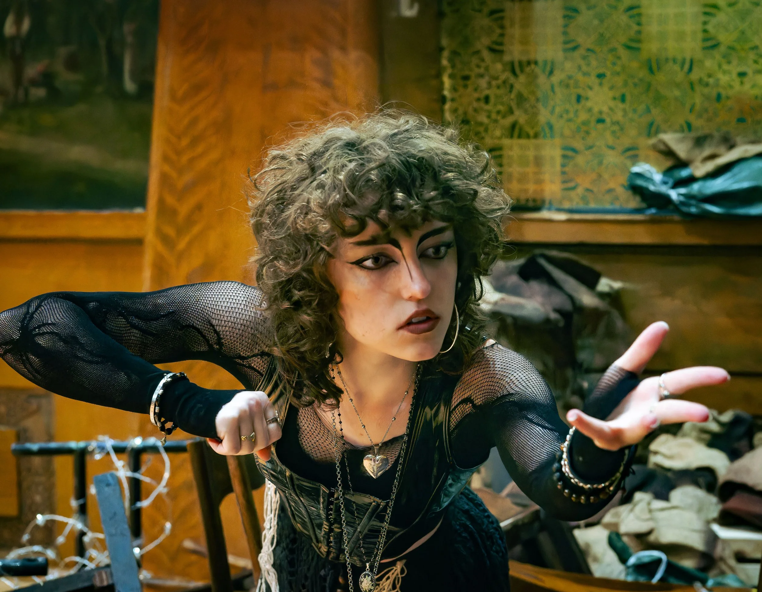 A young singer with curly brown hair and dramatic goth makeup, wearing a black mesh top and layered jewelry, is reaching out with her hand on stage.