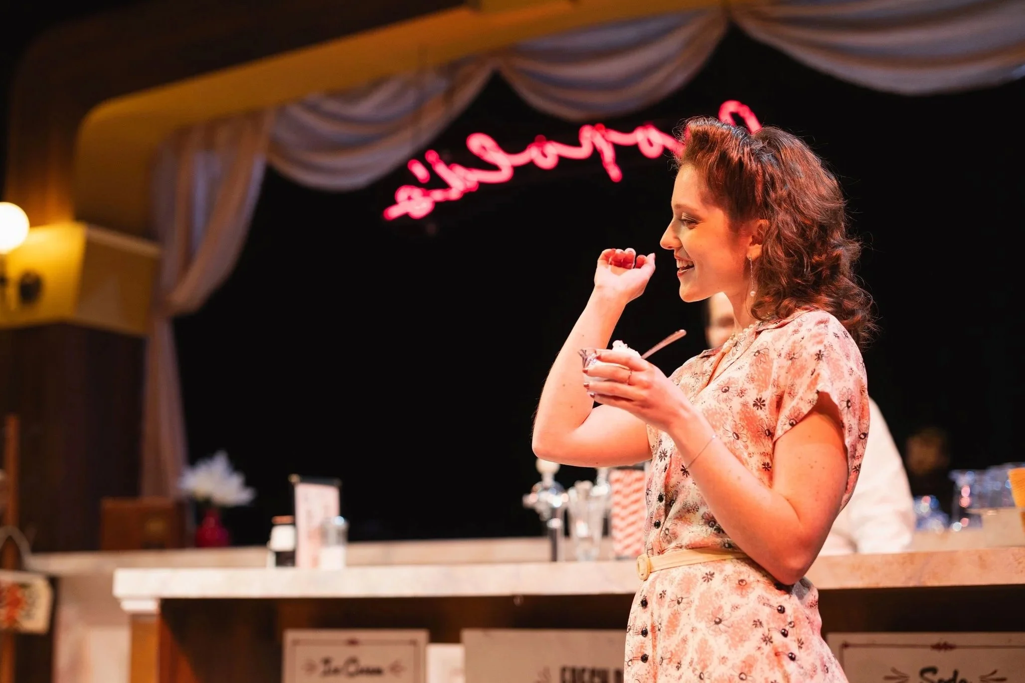 An opera singer with curly brown hair, wearing a floral dress, smiling and holding an ice cream cup, standing in front of an icecream bar on an opera stage with a neon pink sign in the background.