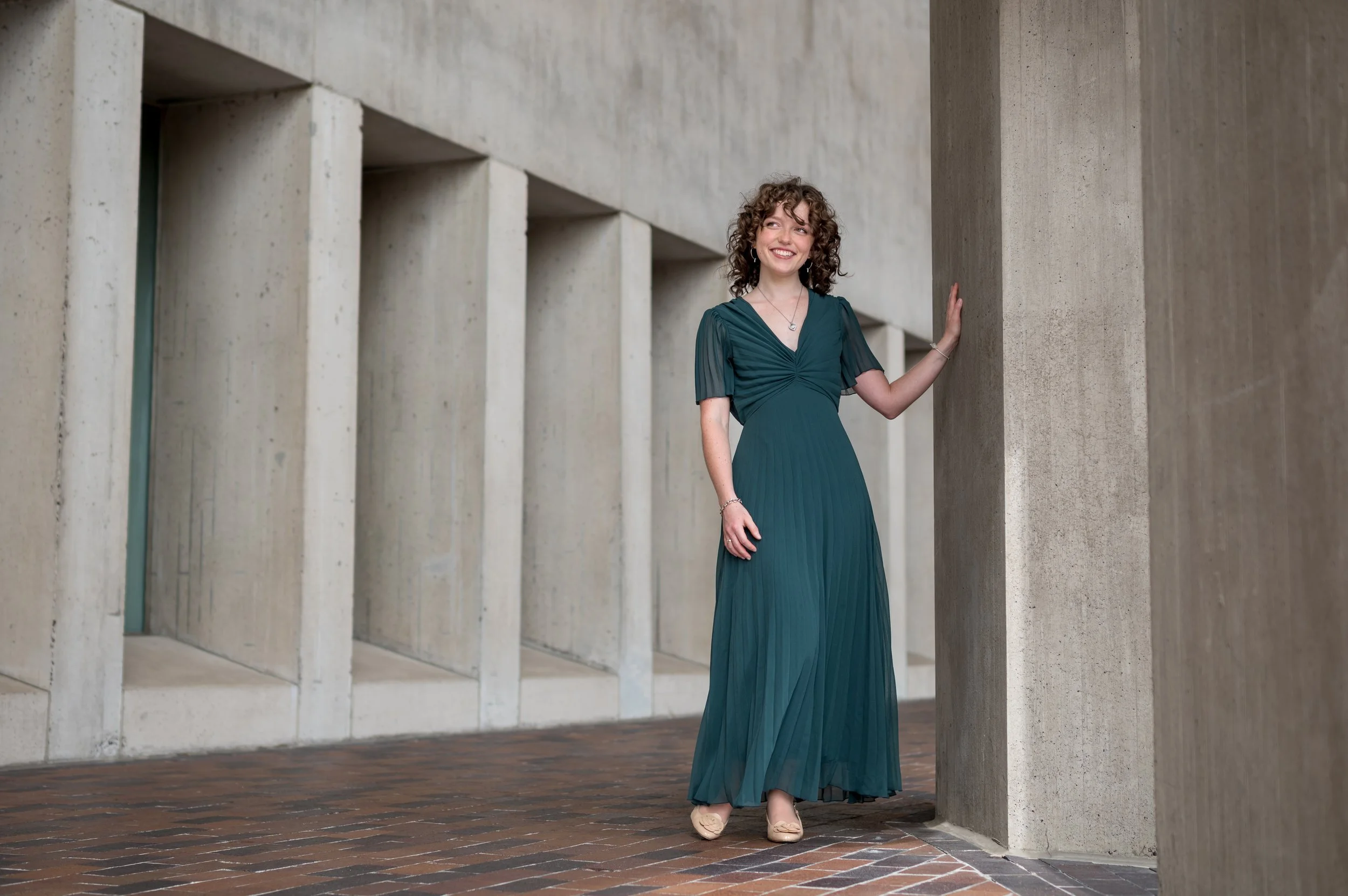 A young woman in a teal dress standing outdoors next to concrete architectural columns, smiling and leaning against a column.