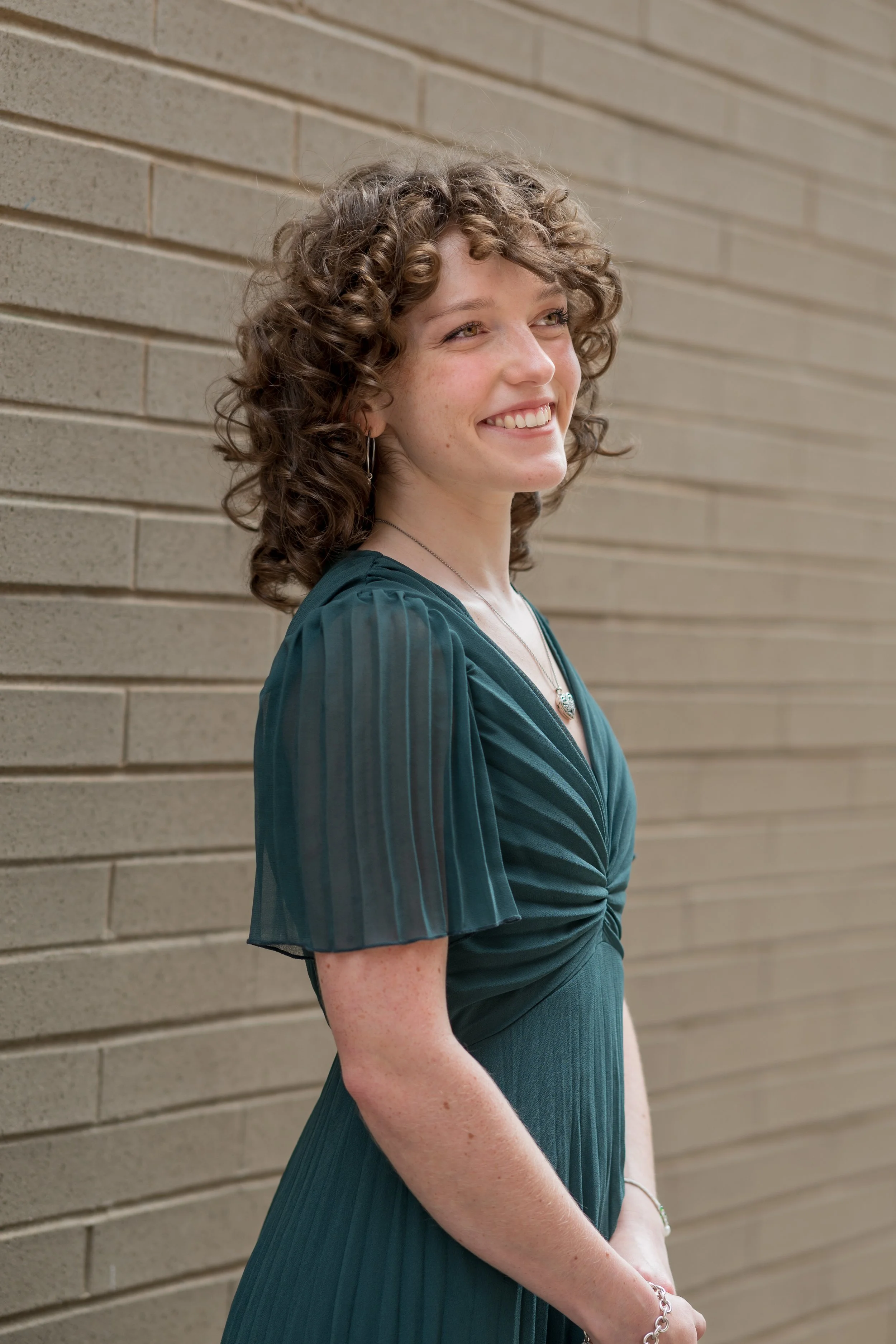A woman with curly brown hair and freckles smiling, standing against a beige brick wall. She is wearing a teal-green dress with pleated, semi-transparent sleeves, a necklace, and a bracelet.