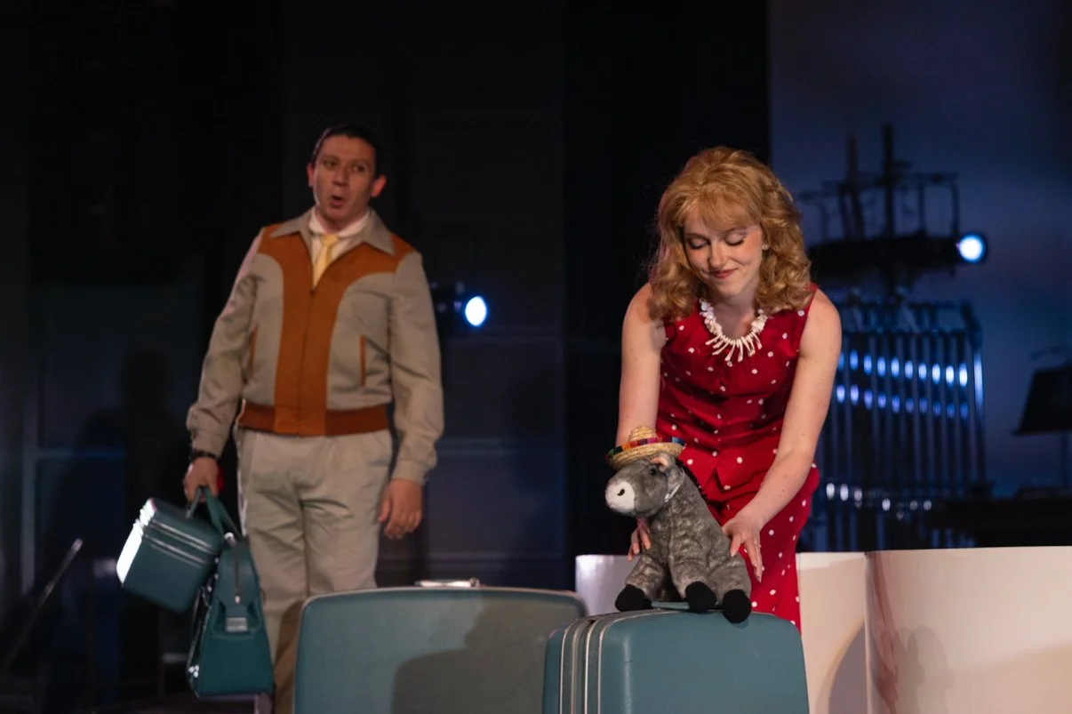 A woman in a red polka dot dress places a stuffed donkey with a sombrero on top on a table. A man in the background holds a teal bag and a watering can, standing in a dimly lit room with blue lighting and stage set pieces.
