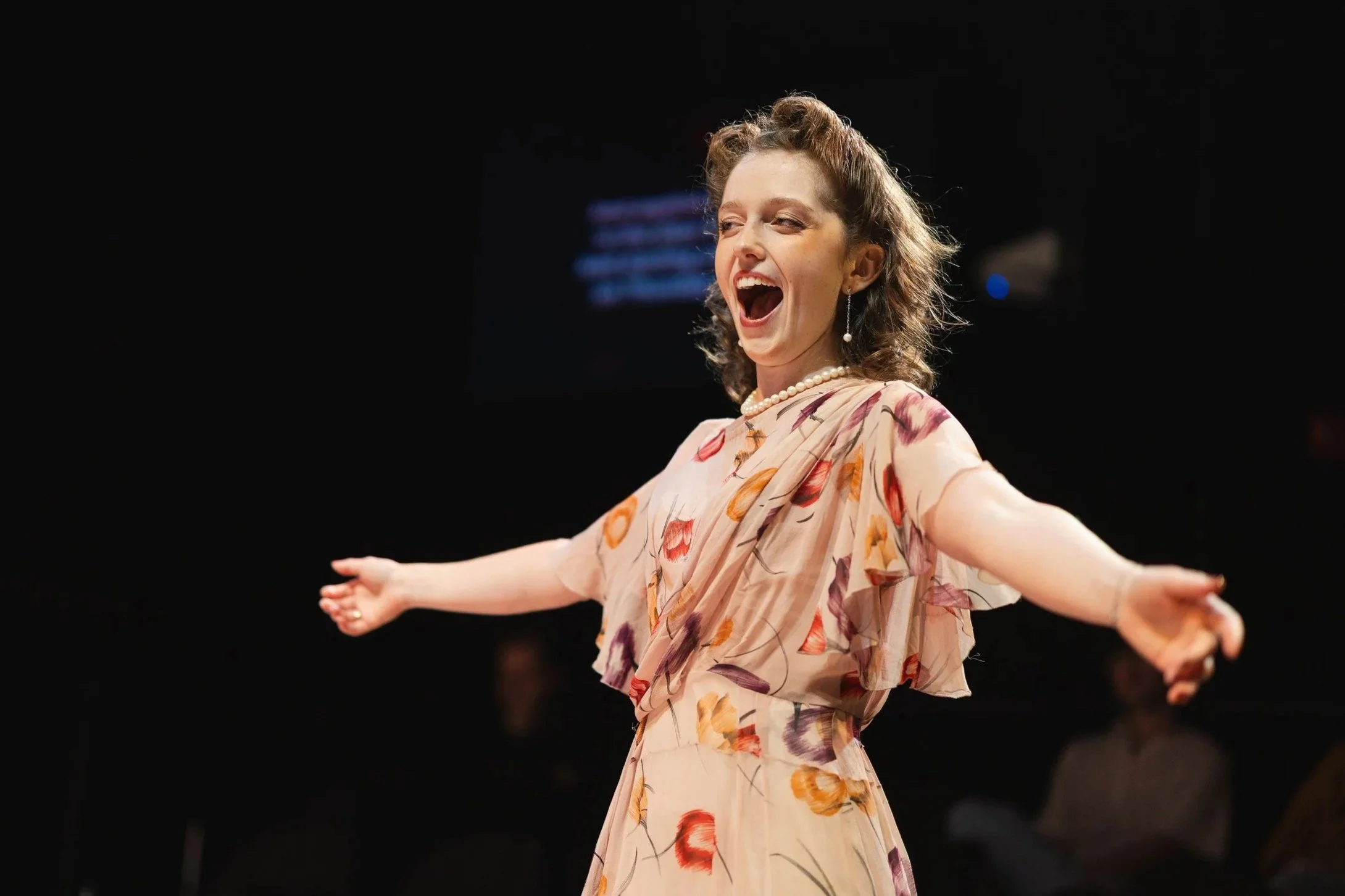 A an opera singer singing with arms outstretched, wearing a floral dress and pearl earrings, on a dark stage.