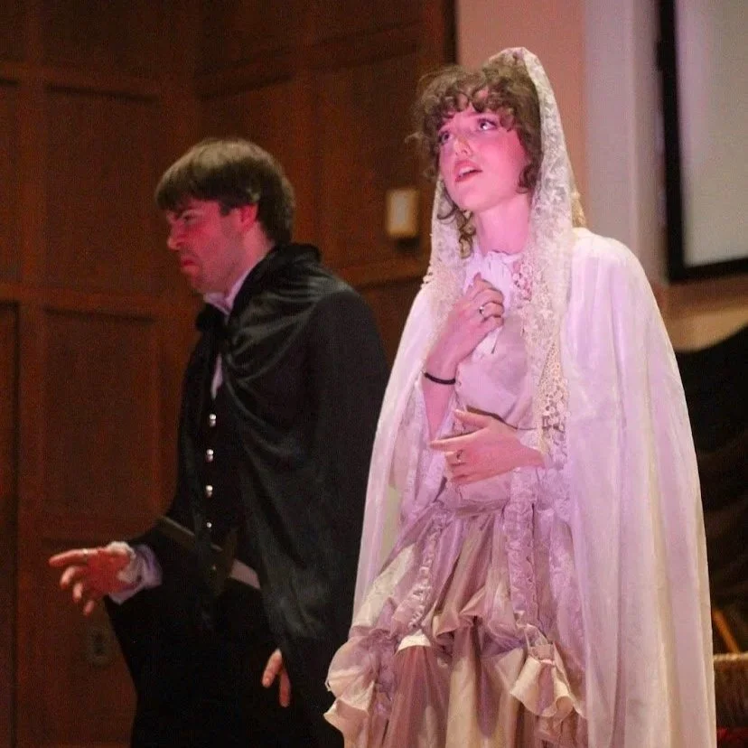 Two opera singers on stage in zarzuela in period costumes; a man dressed as a count and a woman dressed as a princess with a lace hood, appearing emotional or surprised.