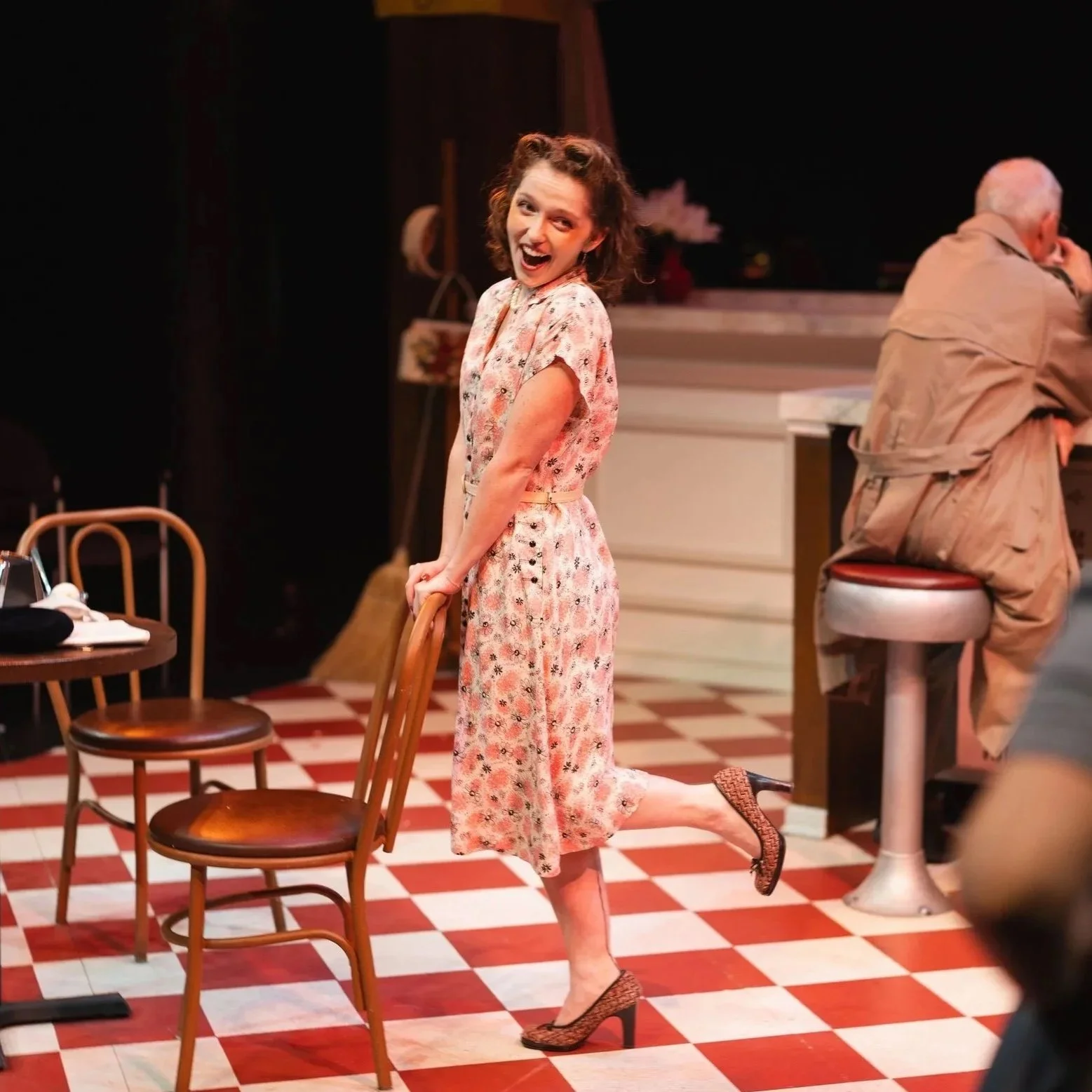 A young woman with curly brown hair, wearing a floral dress and high heels, poses on a checkered floor, smiling in a retro-style set of a contemporary opera.