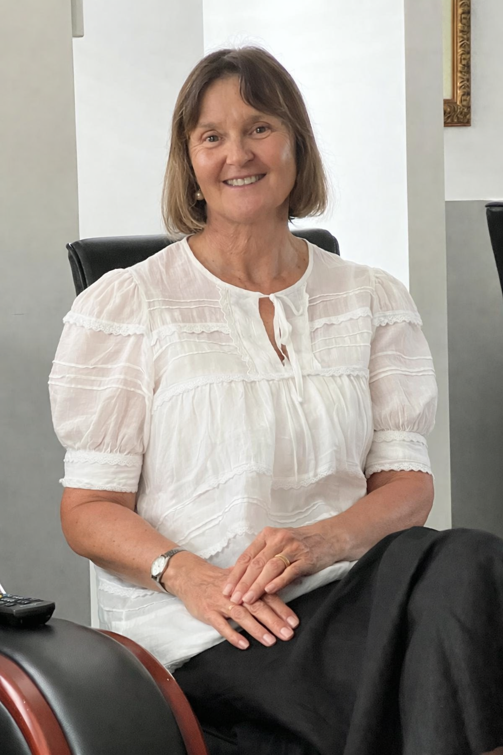 A middle-aged woman with shoulder-length brown hair and light skin, wearing a white blouse with puffed sleeves and a keyhole neckline, seated in a black chair in an indoor setting, smiling at the camera.