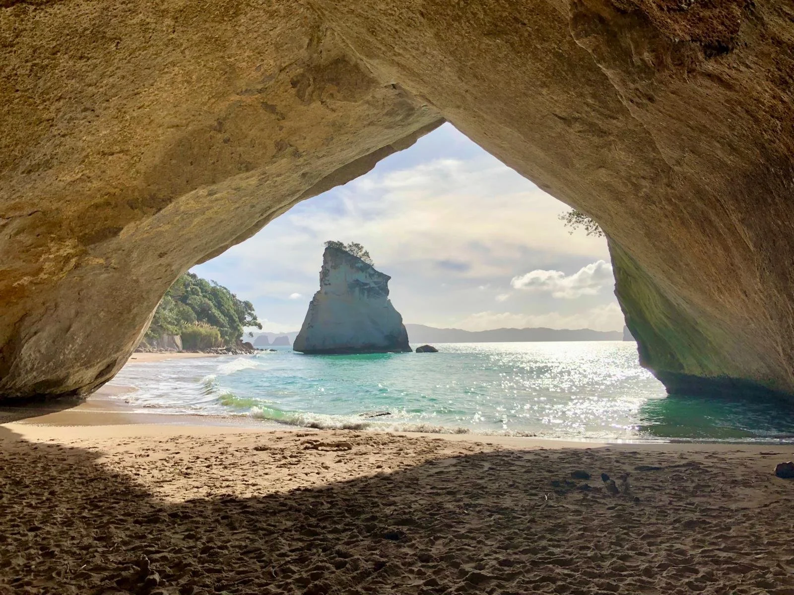 A view of a beach with a large rock formation in the ocean, seen through a natural rock arch. The sky is partly cloudy with bright sunlight reflecting on the water.