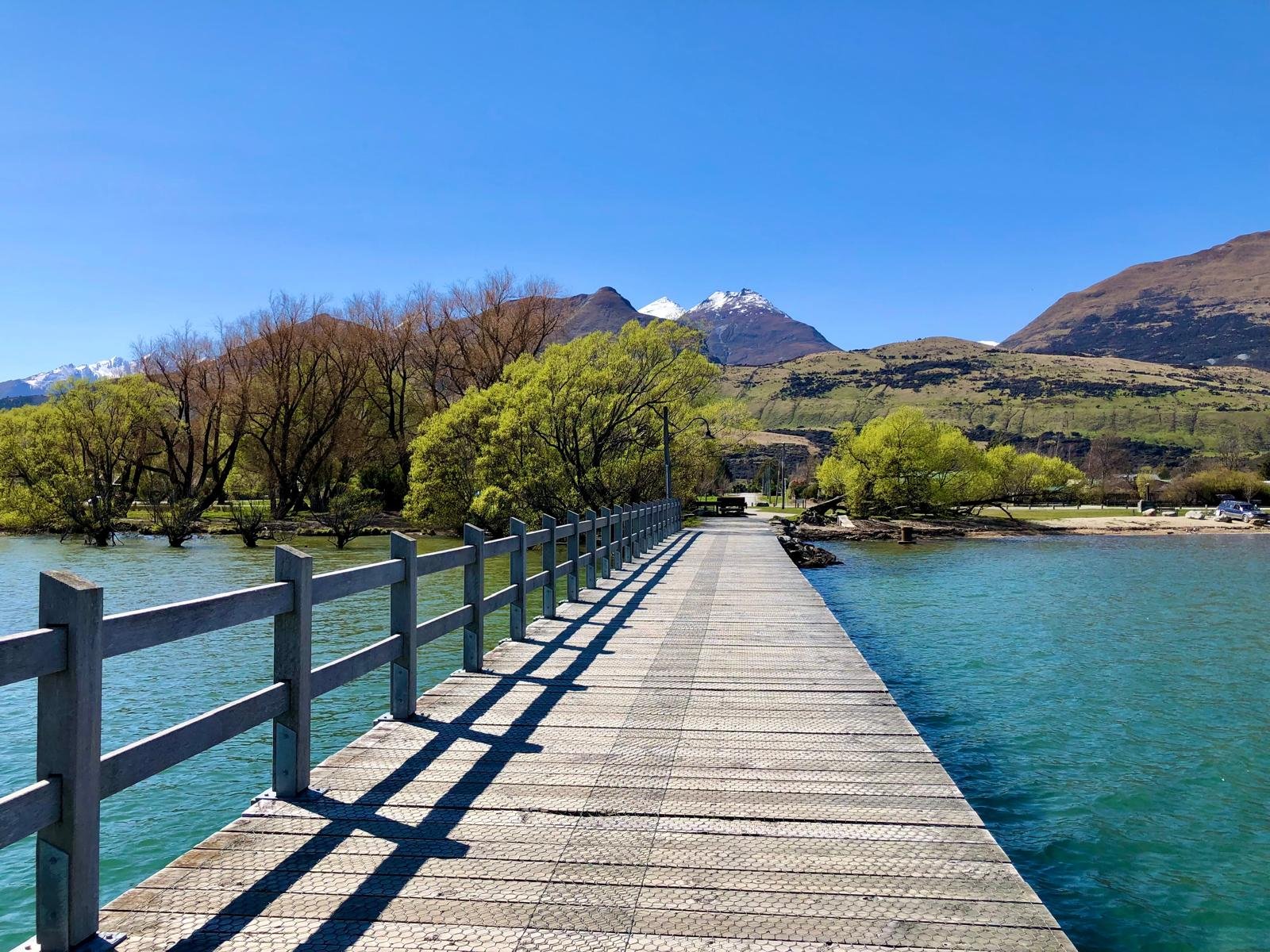 A wooden pier extending over blue water, with a railing on the left, surrounded by trees and mountains with snow on the peaks under a clear blue sky.