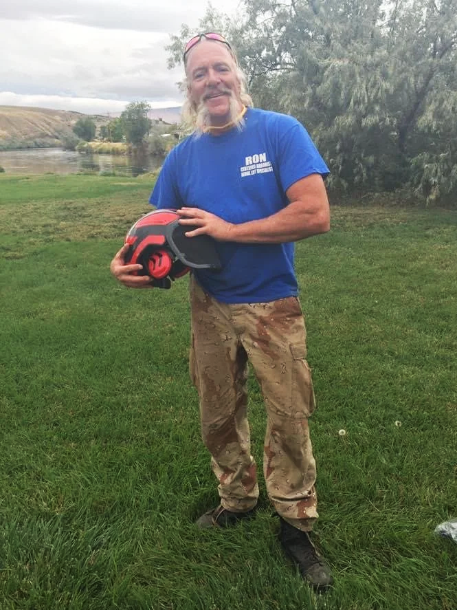 A man with long hair and a mustache smiling outdoors while holding a red and black helmet in his hand, wearing a blue shirt and camouflage pants.