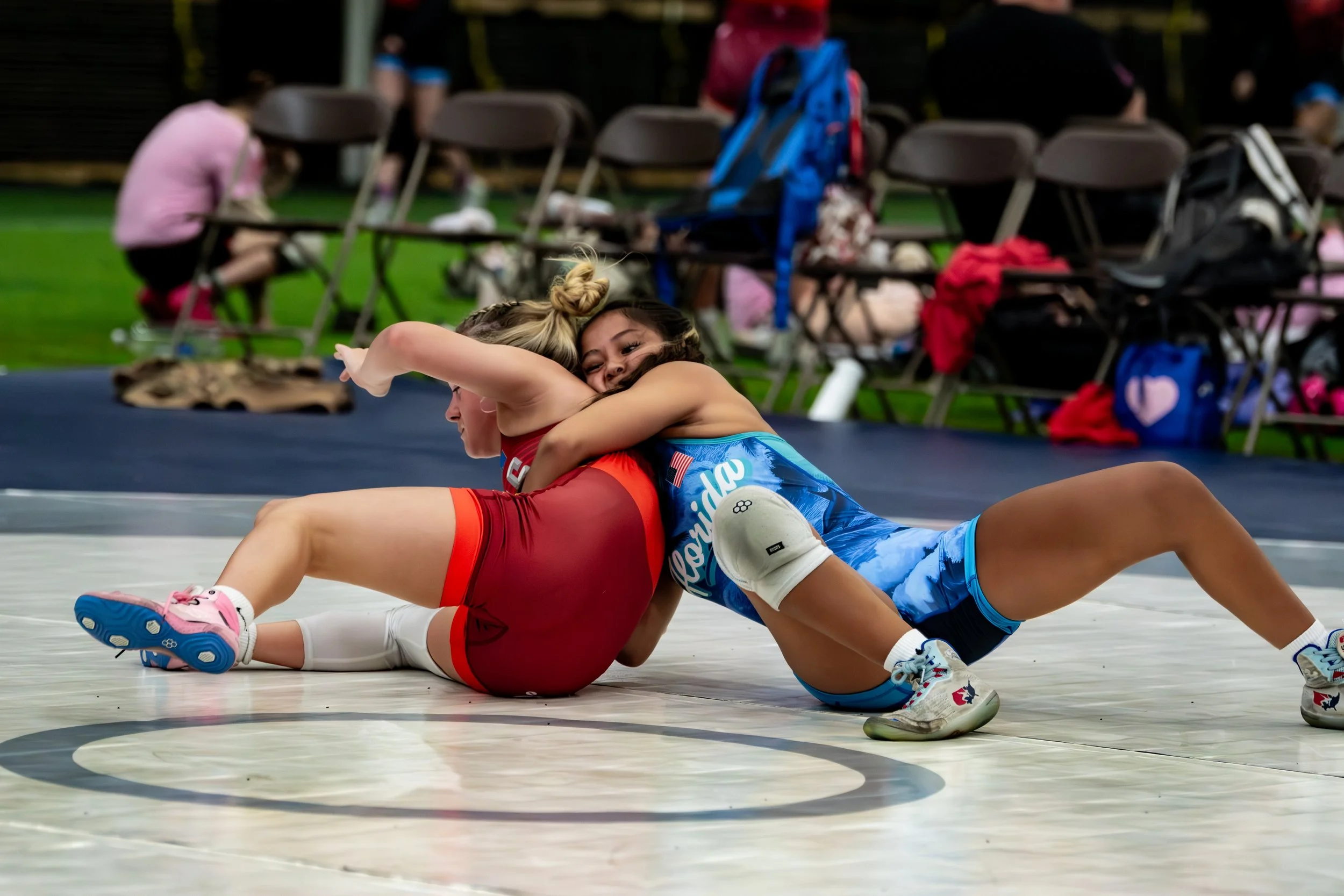 Two female wrestlers in a wrestling match, one in a red and the other in a blue uniform, on a wrestling mat during a competition. They are engaged in a hold, with the wrestler in blue seemingly attempting to pin the opponent in red. The background shows empty chairs and some gear, indicating an indoor sports venue.