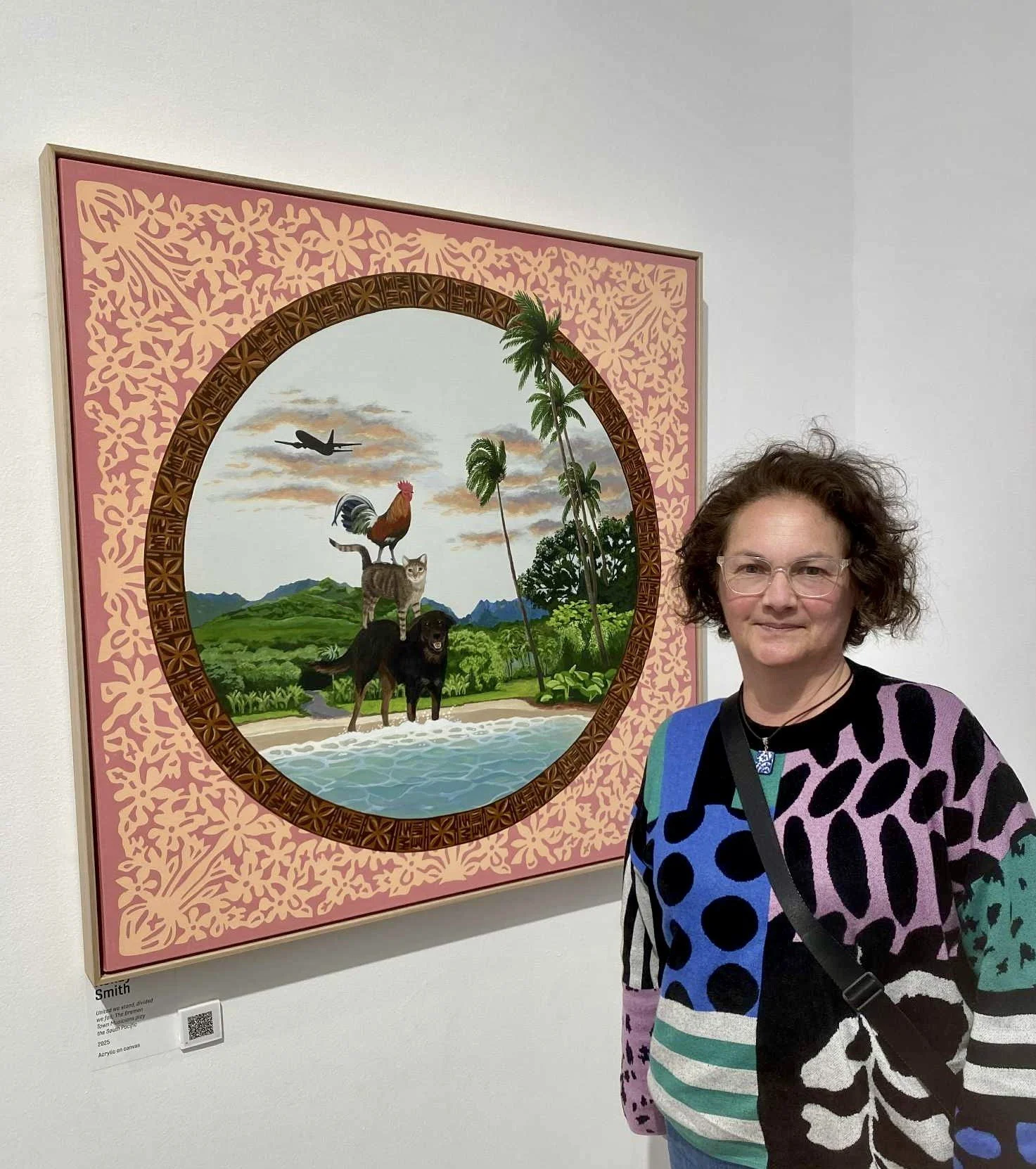 Woman with curly hair and glasses standing in front of a colorful framed painting of nature scene with animals and palm trees.