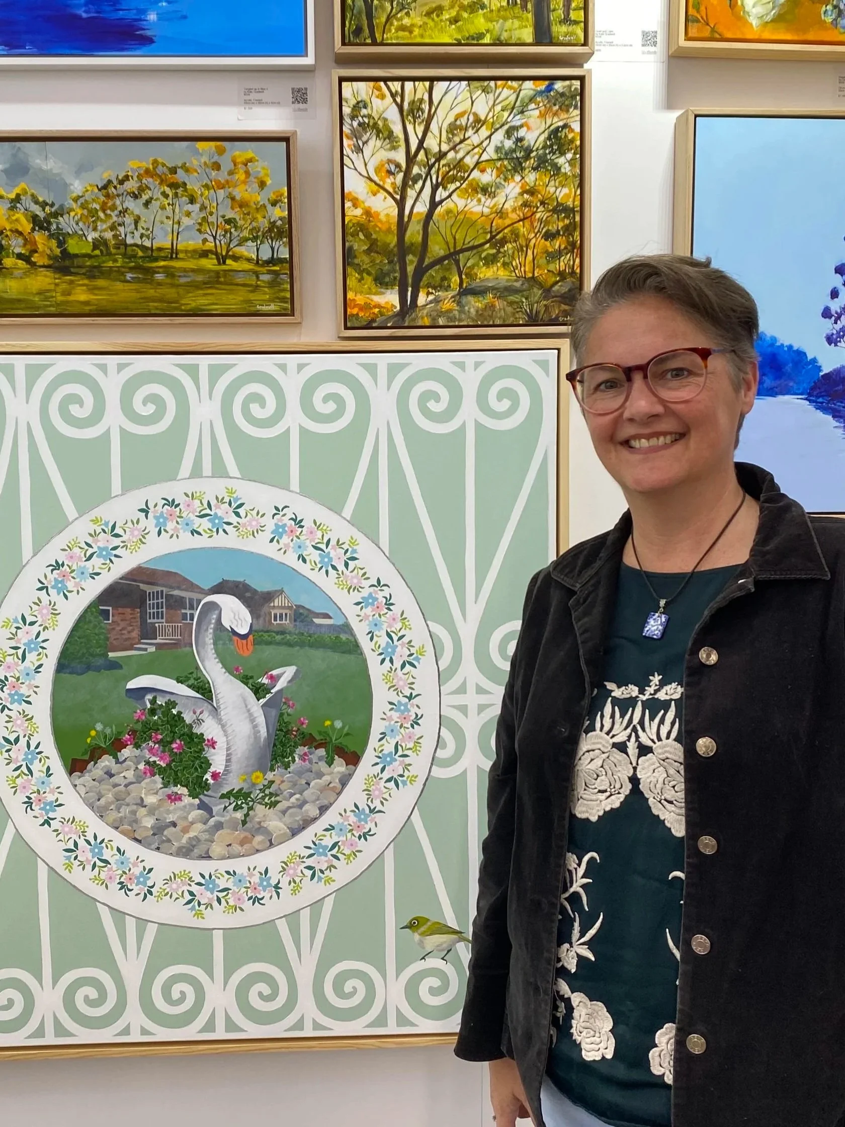 A woman with short gray hair and glasses smiling at an art exhibit. Behind her are several framed landscape paintings of trees and nature scenes. In front of her is a decorative art piece with a swan and flowers.