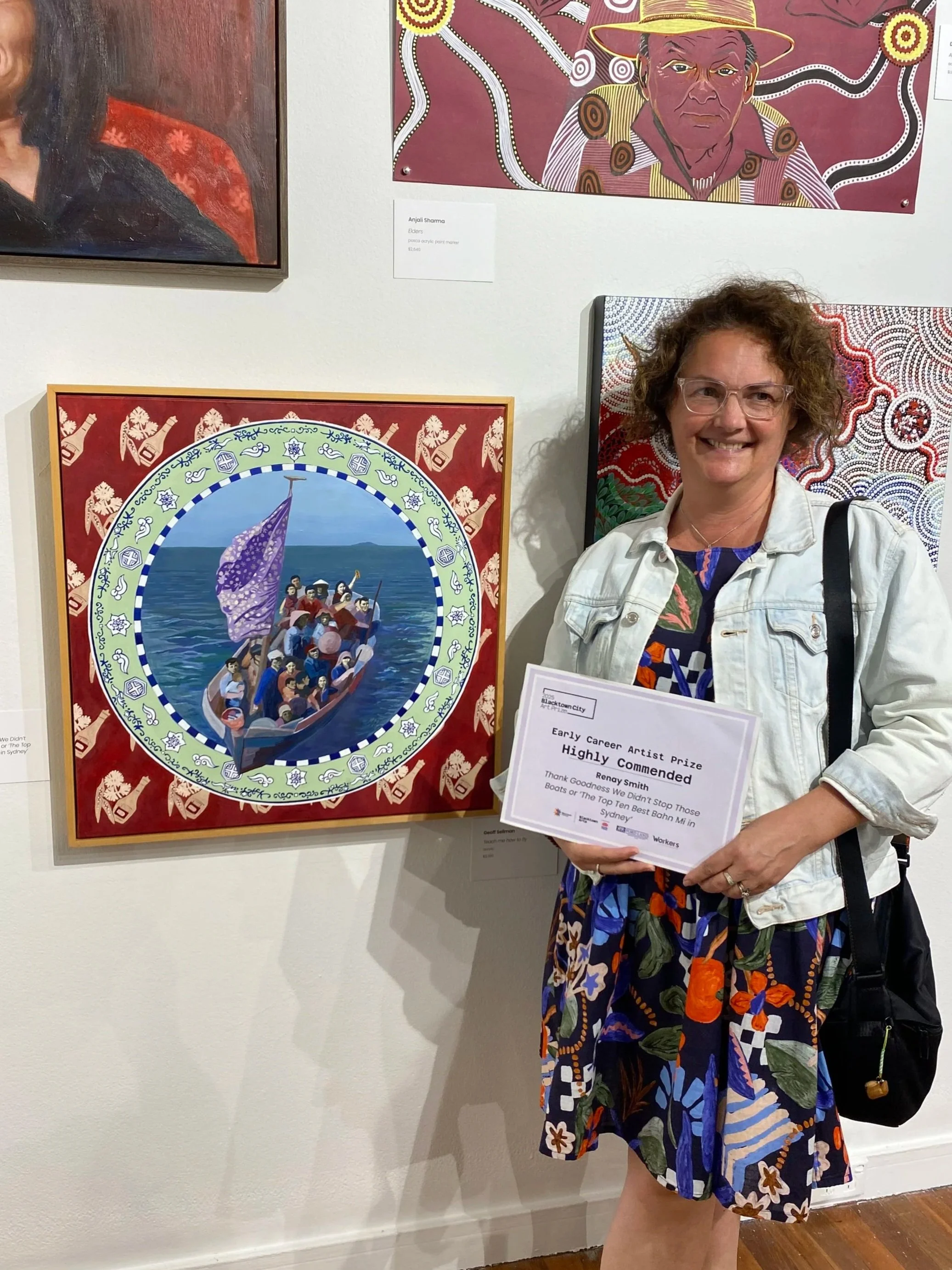 A woman with curly hair and glasses standing in front of colorful artwork, holding a certificate of recognition at an art exhibition.