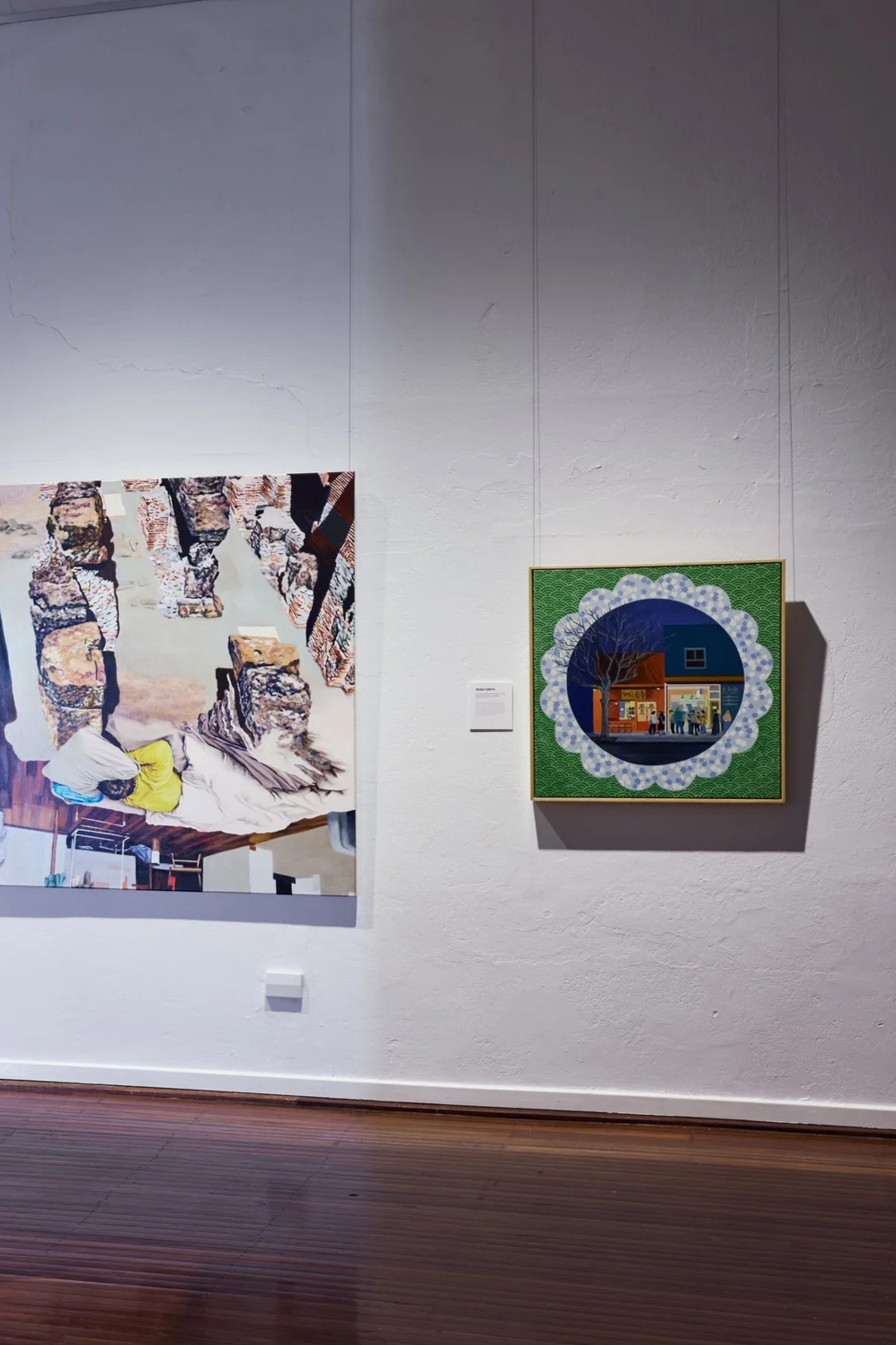 Two paintings on a gallery wall, one featuring a person sleeping among large stones, the other depicting a storefront scene through a decorative circular frame.