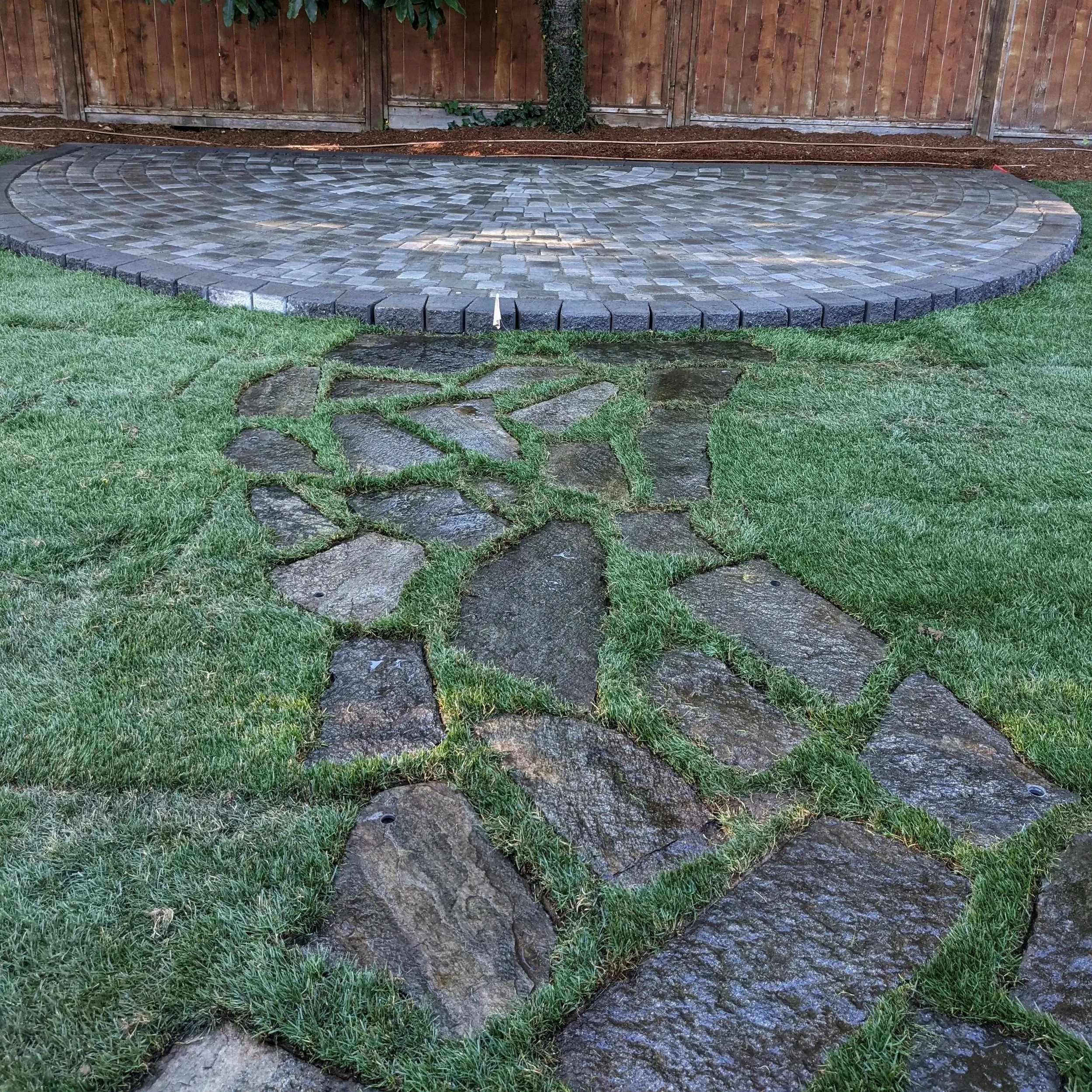 A semi-circular paved backyard patio with green grass and a natural stone pathway leading to the patio