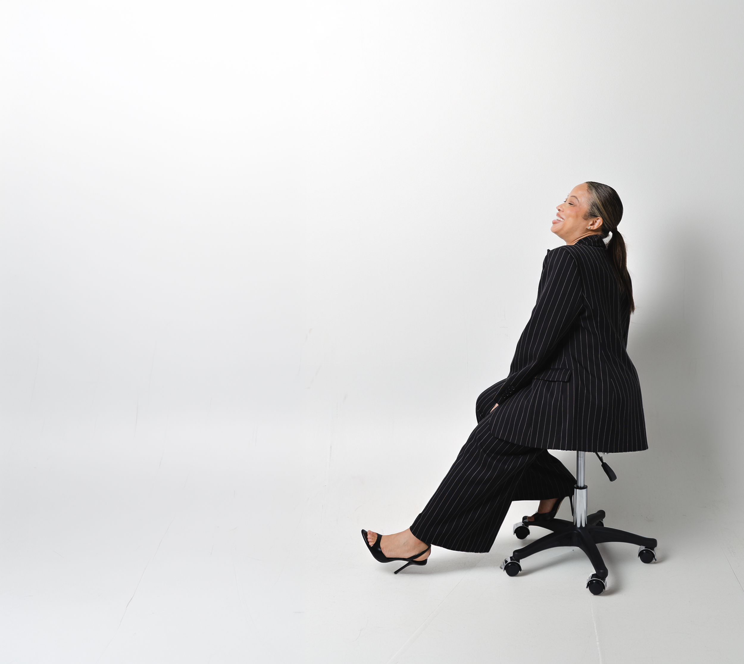 A woman with dark hair tied back, wearing a black pinstripe suit, is sitting on an office chair and smiling with her eyes closed against a plain white background.